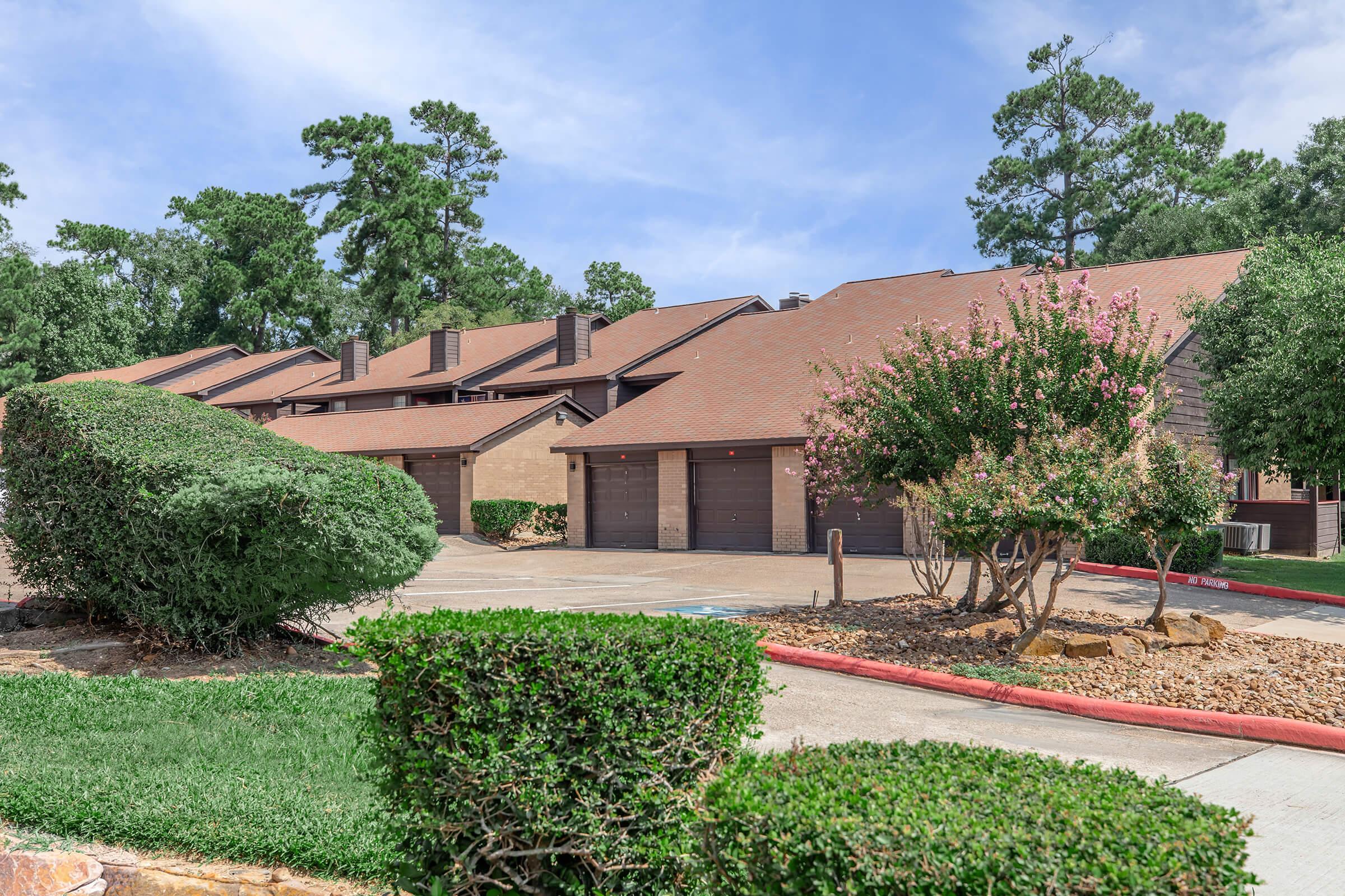 A row of residential buildings featuring brown roofs and attached garages, surrounded by well-maintained lawns and bushes. Lush greenery and flowering plants add to the appeal, with a clear blue sky overhead. The setting exudes a tranquil suburban atmosphere.