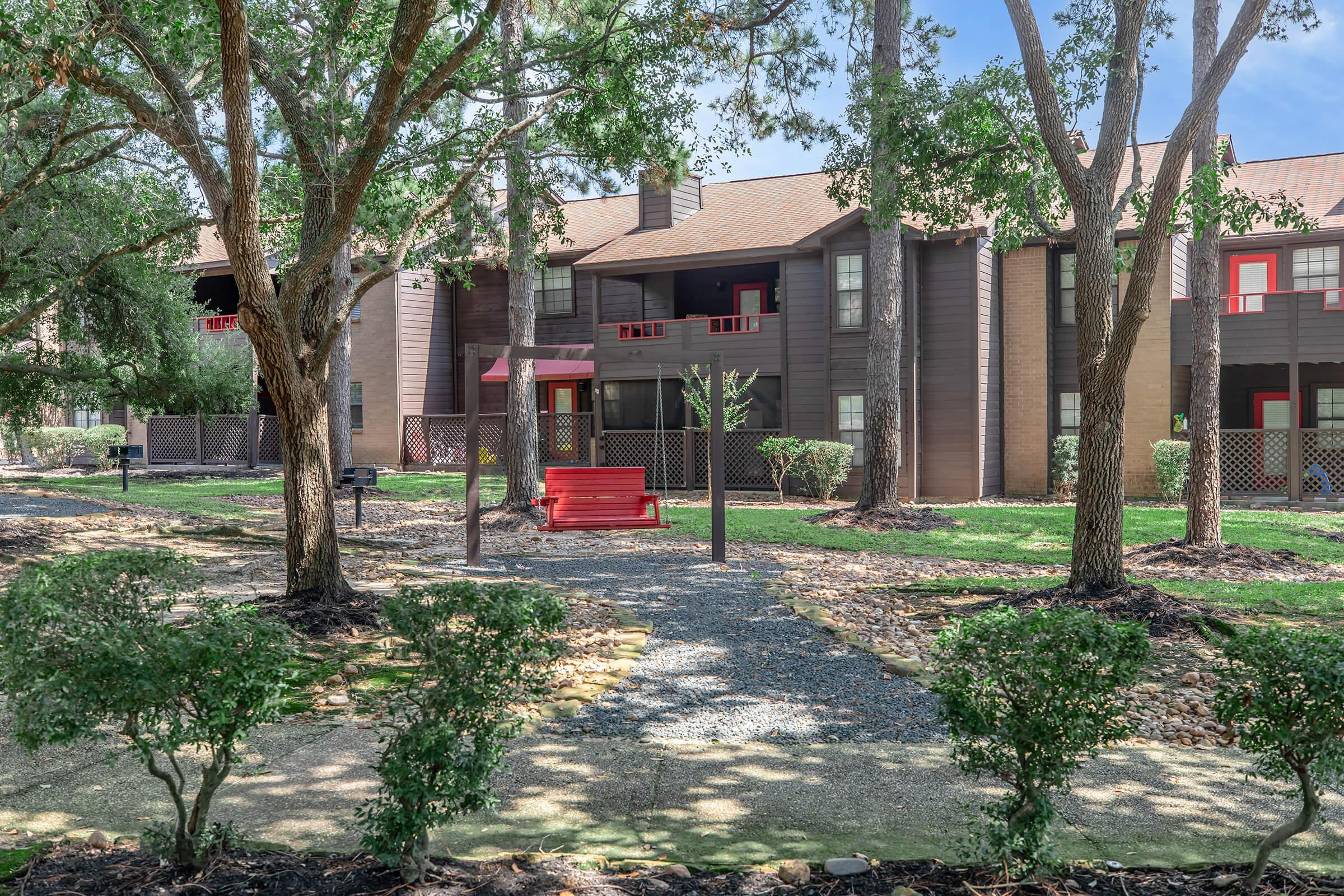 A landscaped area featuring a pathway made of small stones, bordered by green bushes. In the center, a red bench sits under trees, with a multi-story building in the background that has large windows and a mix of dark and light wood siding. The scene is bright and inviting, set in a well-maintained outdoor environment.