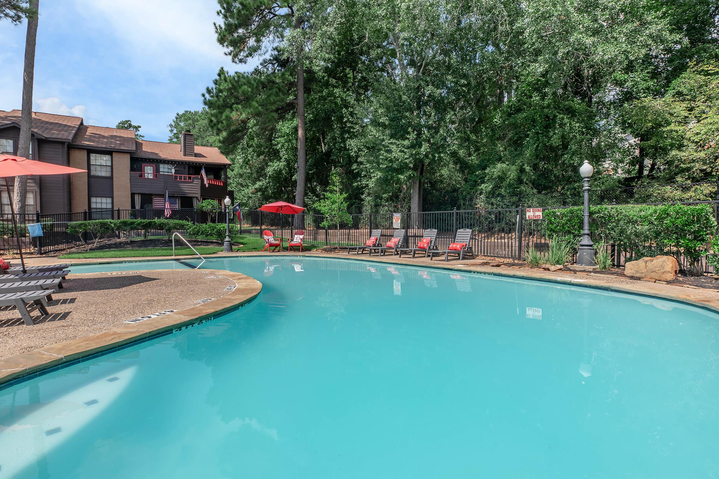 A serene swimming pool surrounded by lounge chairs under red umbrellas, set against a backdrop of lush green trees. The pool area is well-maintained, with a decorative lamp post and a fence for privacy. Perfect for relaxation on a sunny day.