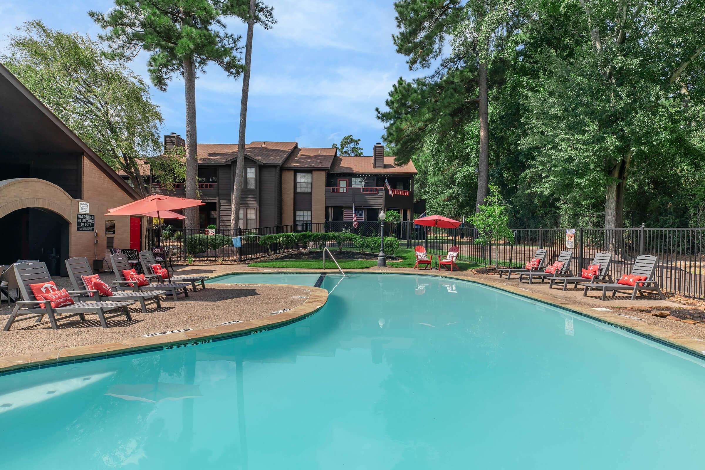 A serene pool area surrounded by green trees and lounge chairs. The pool is sparkling blue, with red umbrellas providing shade. In the background, a multi-story building with balconies is visible, adding to the inviting atmosphere perfect for relaxation and leisure.