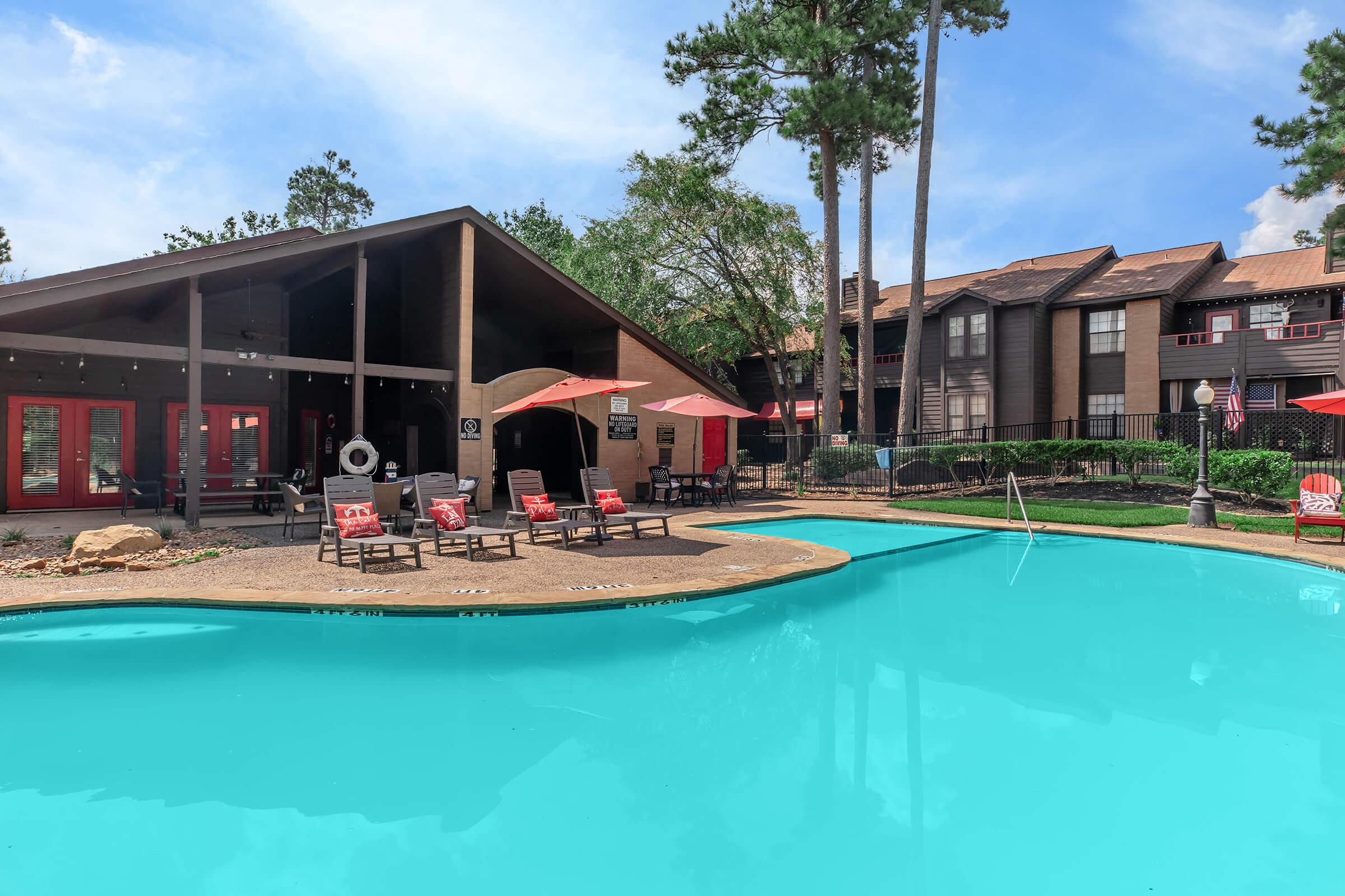 A spacious outdoor pool surrounded by lounge chairs and red umbrellas, with a backdrop of a modern building and tall trees under a clear blue sky. The pool's vibrant turquoise water reflects the sunlight, creating an inviting atmosphere for relaxation.