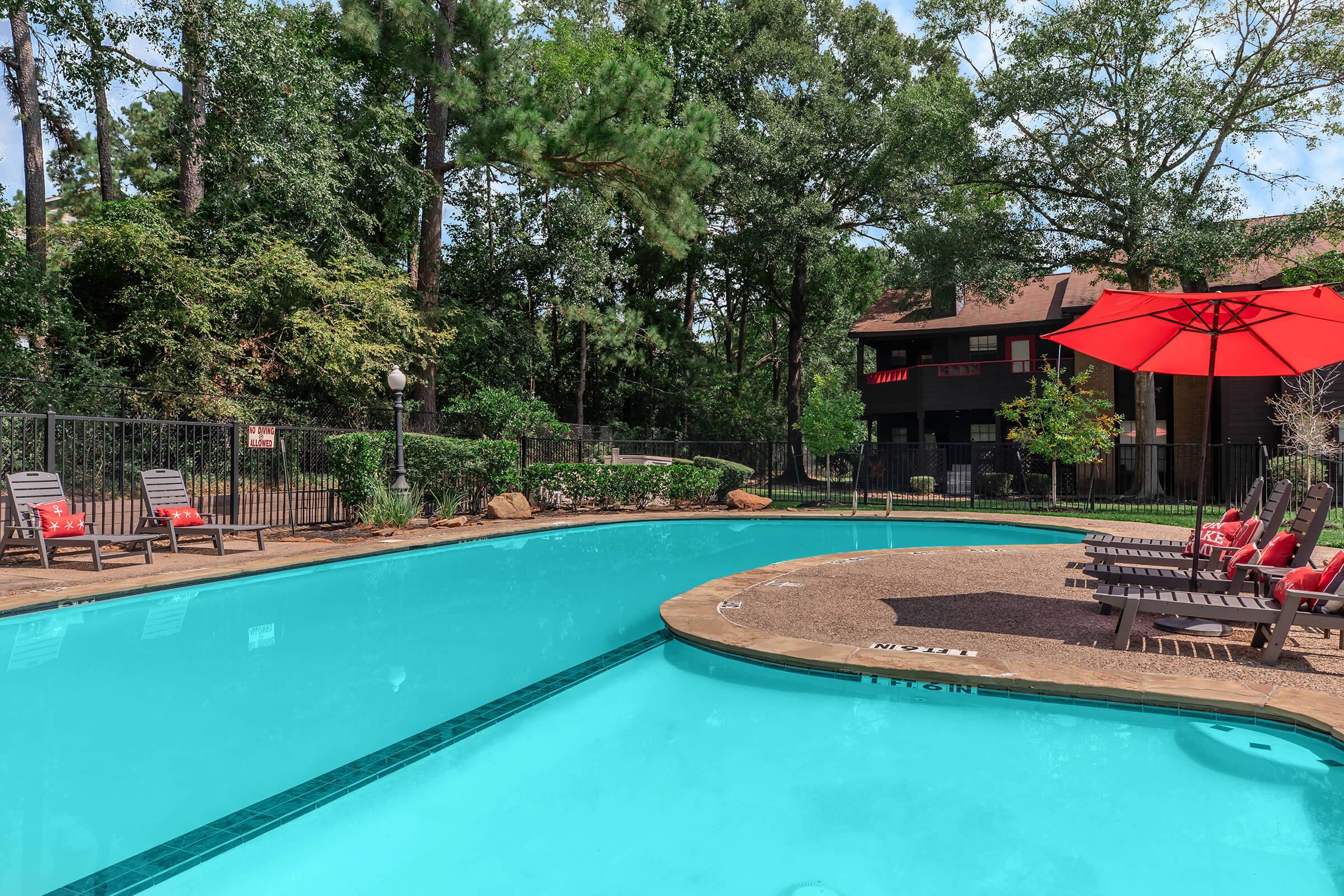 A serene outdoor swimming pool surrounded by greenery, featuring lounge chairs and a red umbrella. The pool's blue water reflects the sky, while a nearby building is partially visible through the trees, adding to the tranquil atmosphere.