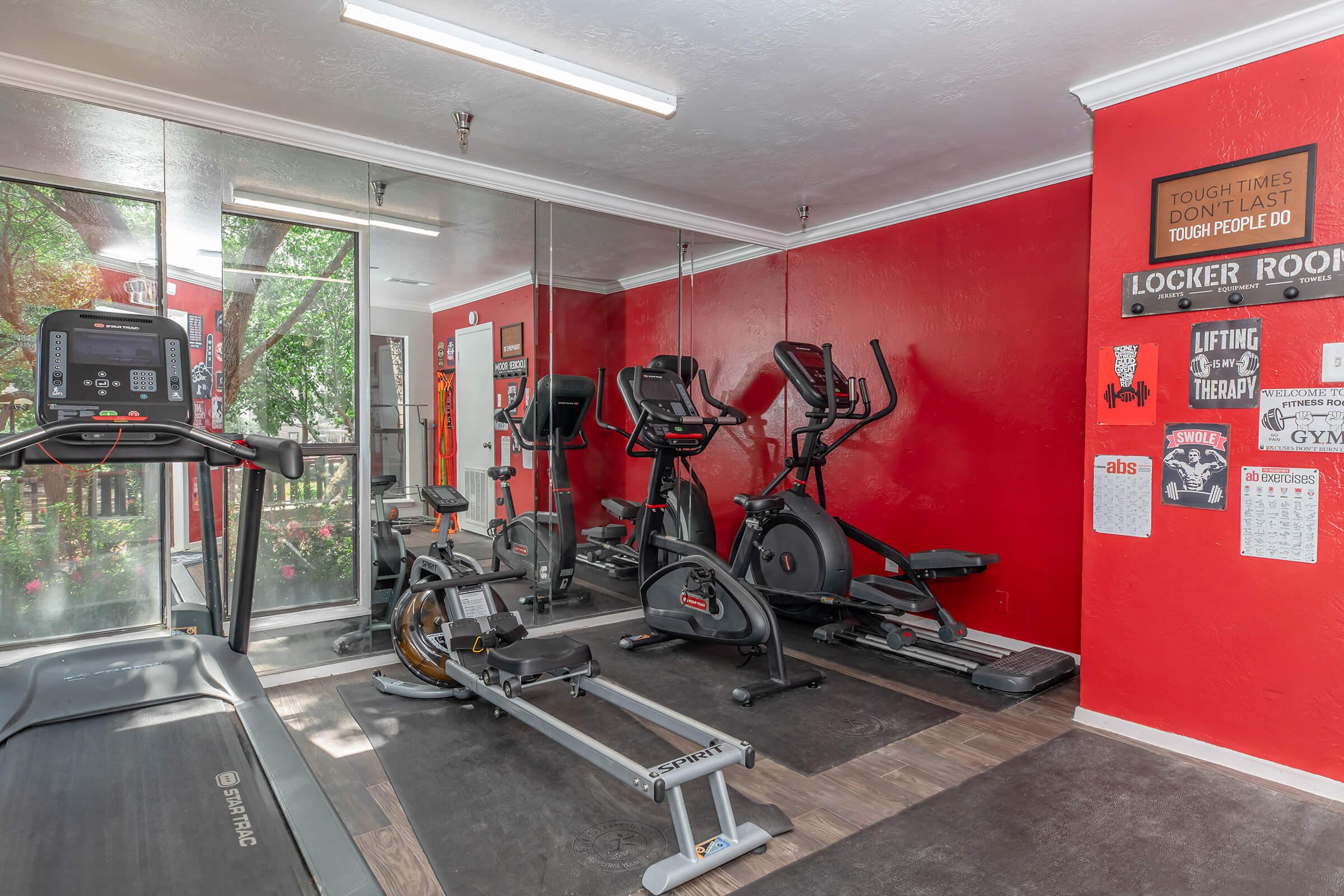Interior of a gym featuring a bright red wall. Equipment includes an elliptical machine, a rowing machine, and a treadmill. Large windows provide natural light, and there are fitness-related posters on the wall. A locker room door is visible in the background, adding to the gym atmosphere.