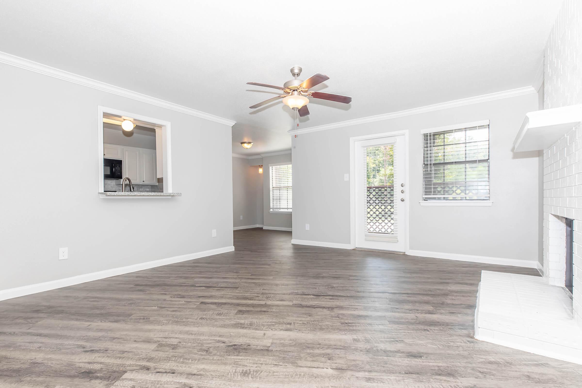 A spacious living room featuring light gray walls and laminate flooring. It has a ceiling fan and a fireplace with a white surround. There's a doorway leading to a small balcony, and a window allowing natural light. The room is bright and inviting with a modern aesthetic.