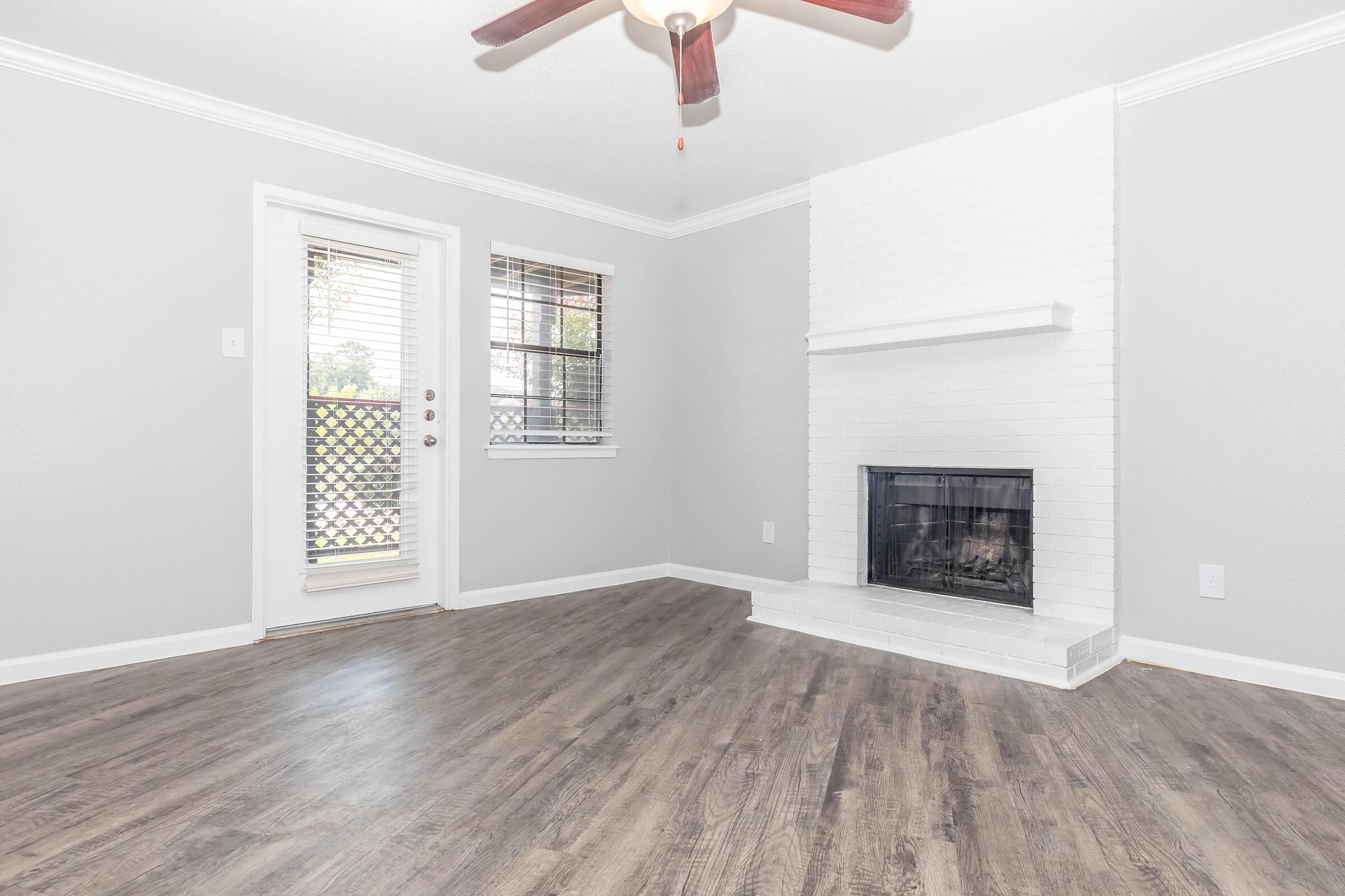 A brightly lit living room featuring light gray walls, a white brick fireplace, and wooden flooring. There is a ceiling fan with wooden blades, and a door leading to an outdoor space is visible beside a window with blinds. The room has a clean and modern aesthetic.