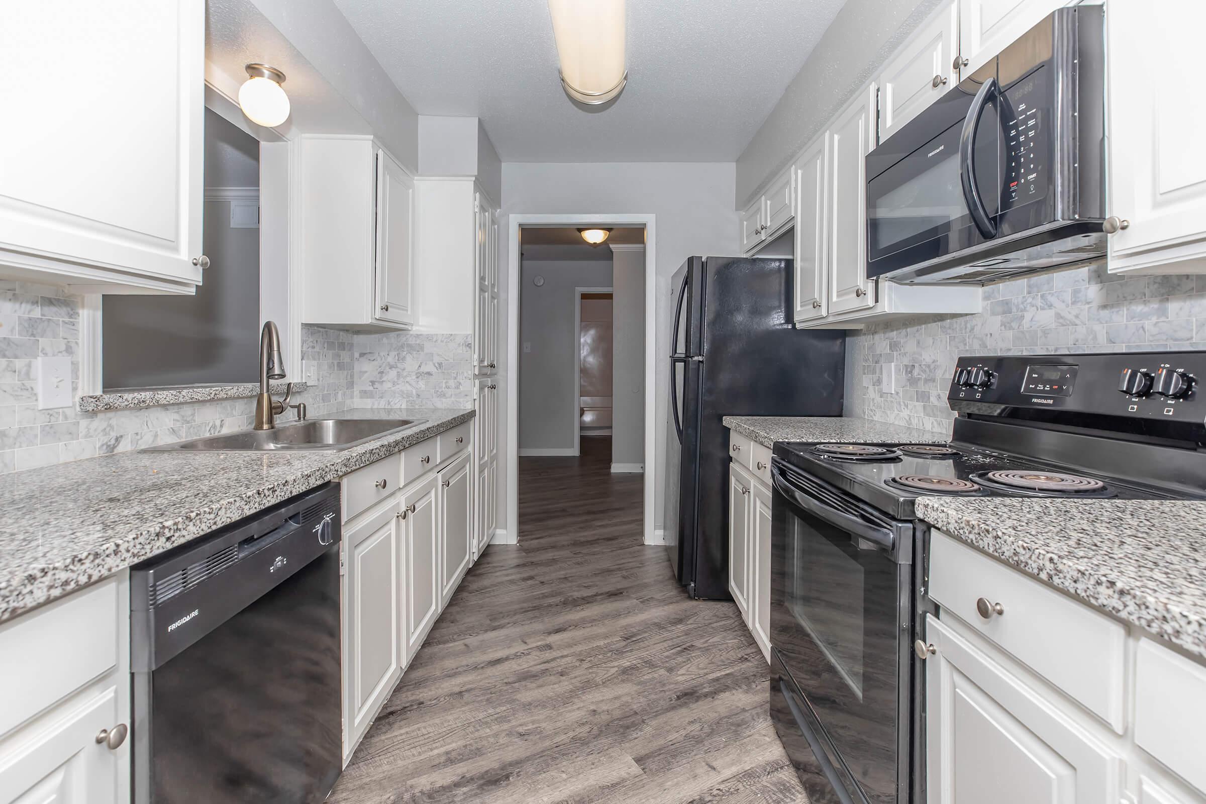 A modern kitchen featuring white cabinets, granite countertops, stainless steel sink, and appliances including a microwave, stove, and refrigerator. The flooring is a dark wood-like material, and the space is well-lit with an overhead light, leading to an adjacent room.