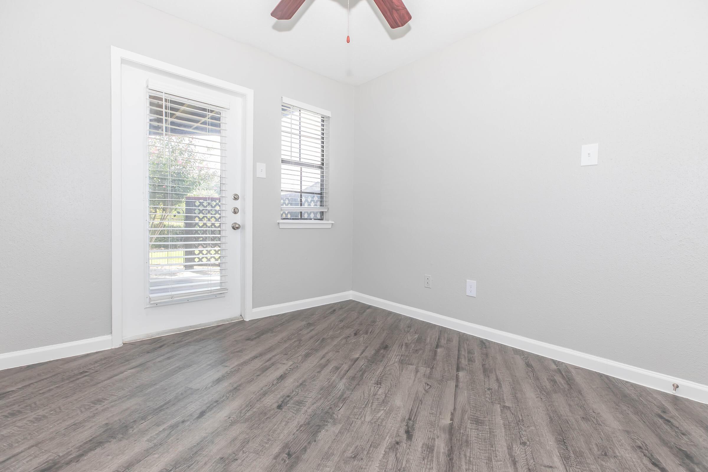 A small, empty room with light gray walls and a wooden floor. It features a ceiling fan and two windows, one with blinds. A door leads outside, allowing natural light to fill the space. The overall look is modern and airy, suitable for various uses such as a living area or office.