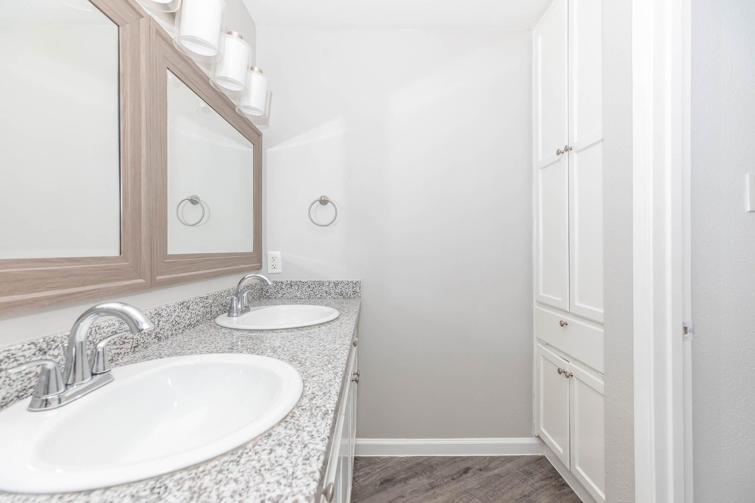A modern bathroom featuring dual sinks with white countertops, large mirrors above each sink, and stylish lighting fixtures. The walls are painted in a light color, and there are white cabinets and a towel ring on the wall. The floor has a wood-like finish.