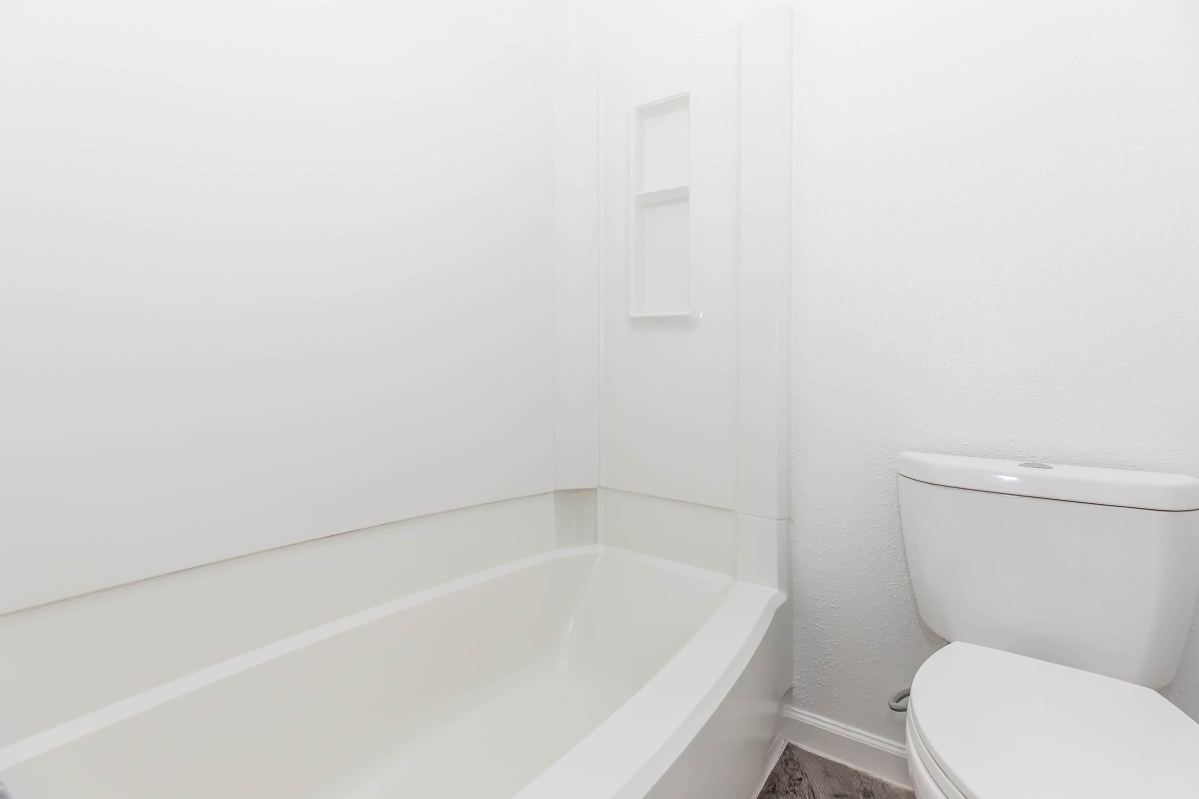 A clean, modern bathroom featuring a white bathtub and a toilet. The walls are painted in light colors, and there is a small shelf nook embedded in the wall above the tub. The floor is tiled, adding a sleek look to the space. Natural light enhances the bright ambiance.