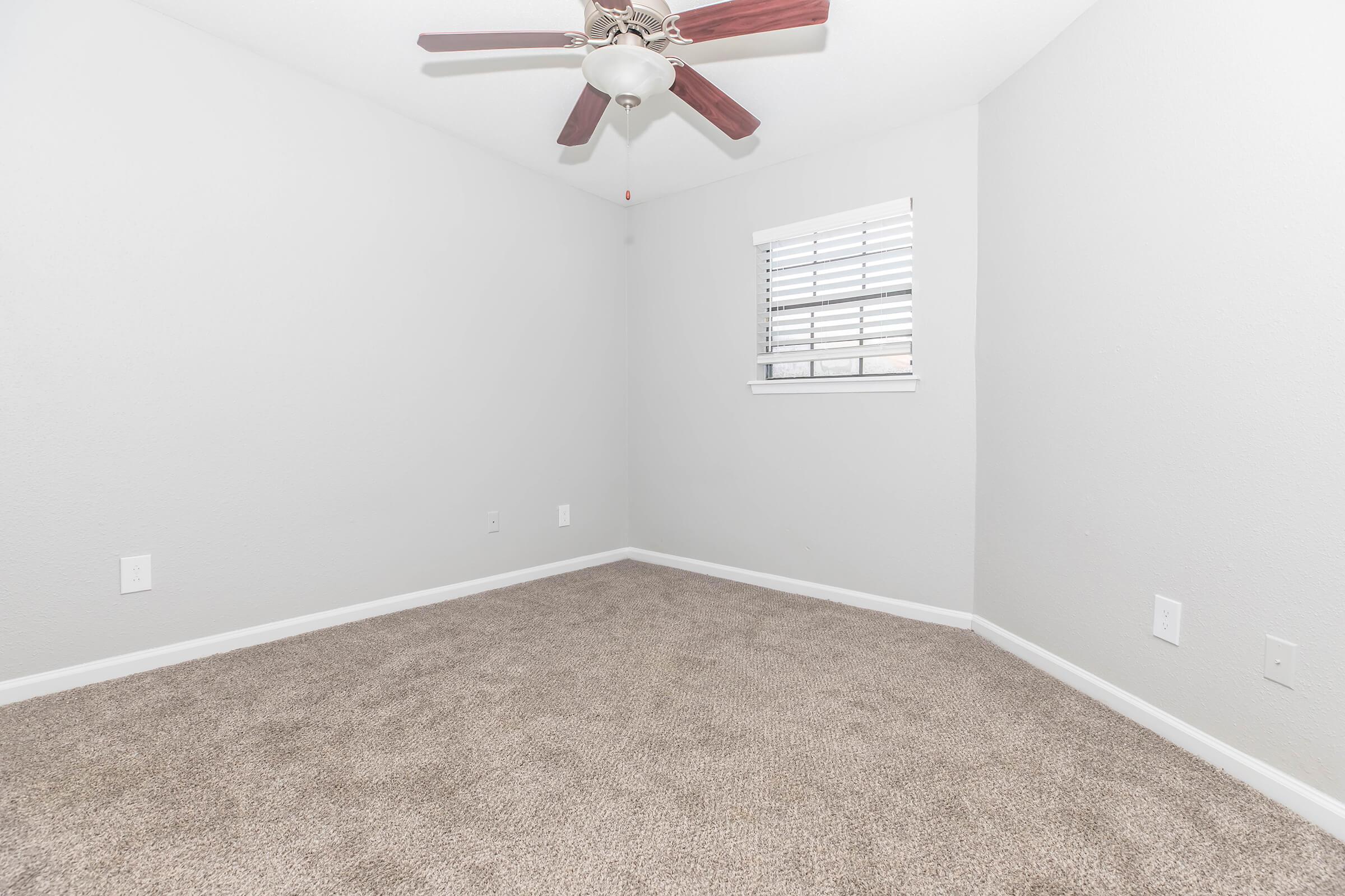 A spacious, empty room with light gray walls and tan carpet. A ceiling fan with wooden blades hangs from the ceiling, and there's a small window with white shutters on one wall. The room has no furniture, giving a clean and open feel.