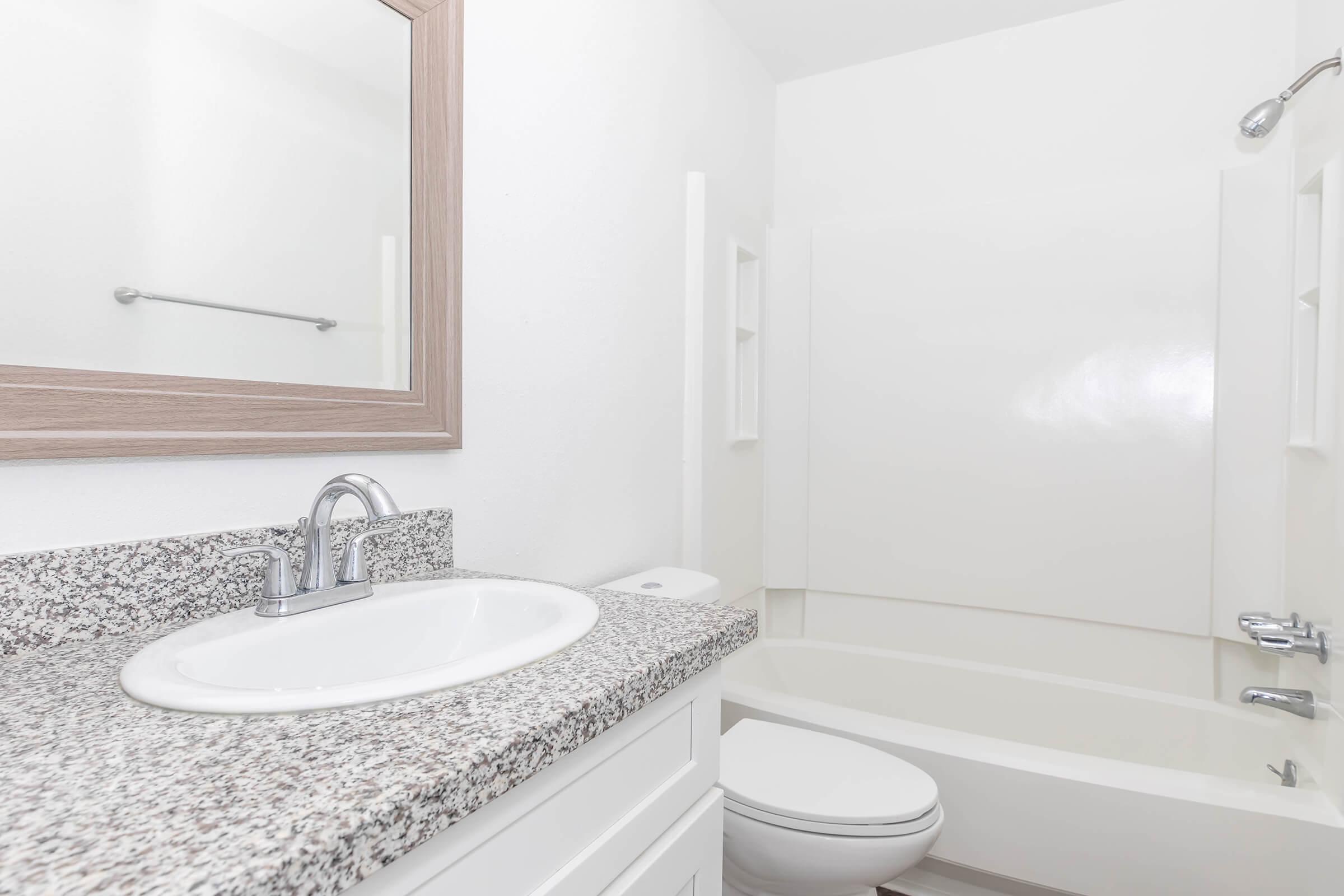A clean and modern bathroom featuring a granite countertop with a sink, a mirror above it, a white bathtub with a shower, and a toilet. The walls are painted white, providing a bright and spacious appearance.