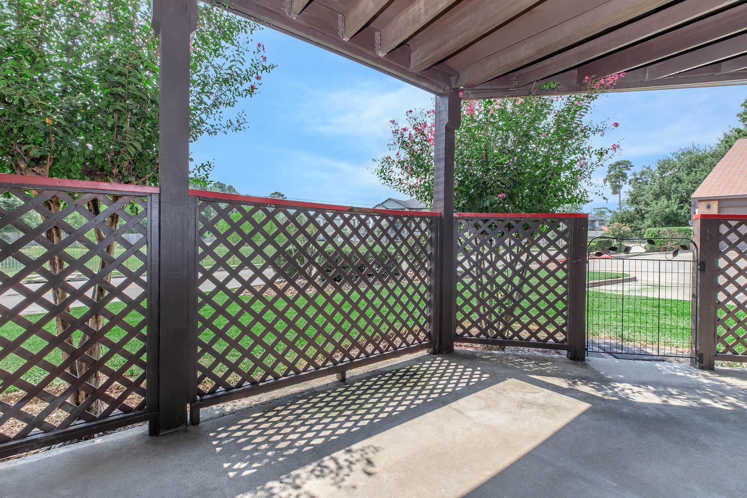 A shaded outdoor patio featuring a wooden lattice fence, with greenery and a clear blue sky visible in the background. The concrete floor is bathed in sunlight, creating a warm and inviting atmosphere.