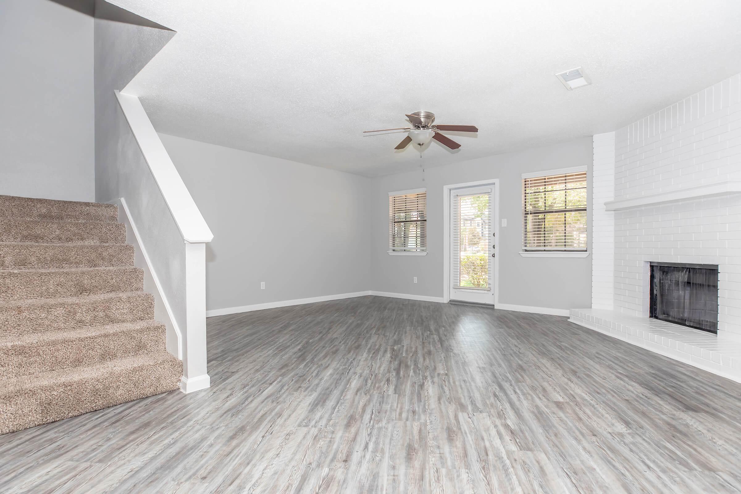 Spacious living room with light gray walls, a ceiling fan, and hardwood-style flooring. A large window lets in natural light, and there's a fireplace on one side. A staircase with beige carpeting leads to the upper level, and a glass door opens to an outdoor area.