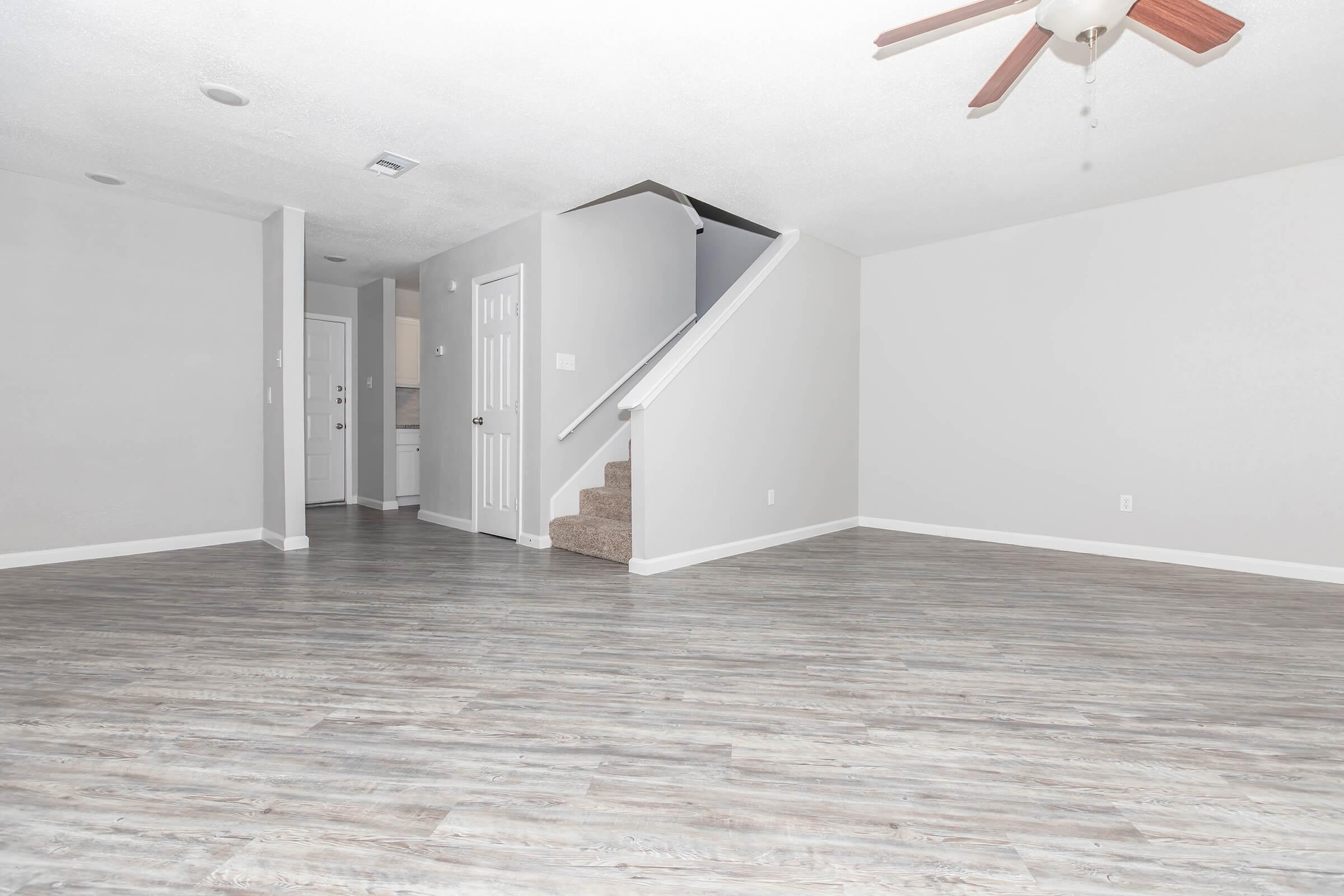 A spacious, empty living area with light gray walls and a ceiling fan, featuring a modern wood-look floor. A staircase leads to an upper level on the right, and a doorway in the back opens to a kitchen or hallway area. The overall ambiance is clean and bright, ideal for potential furnishings and decor.