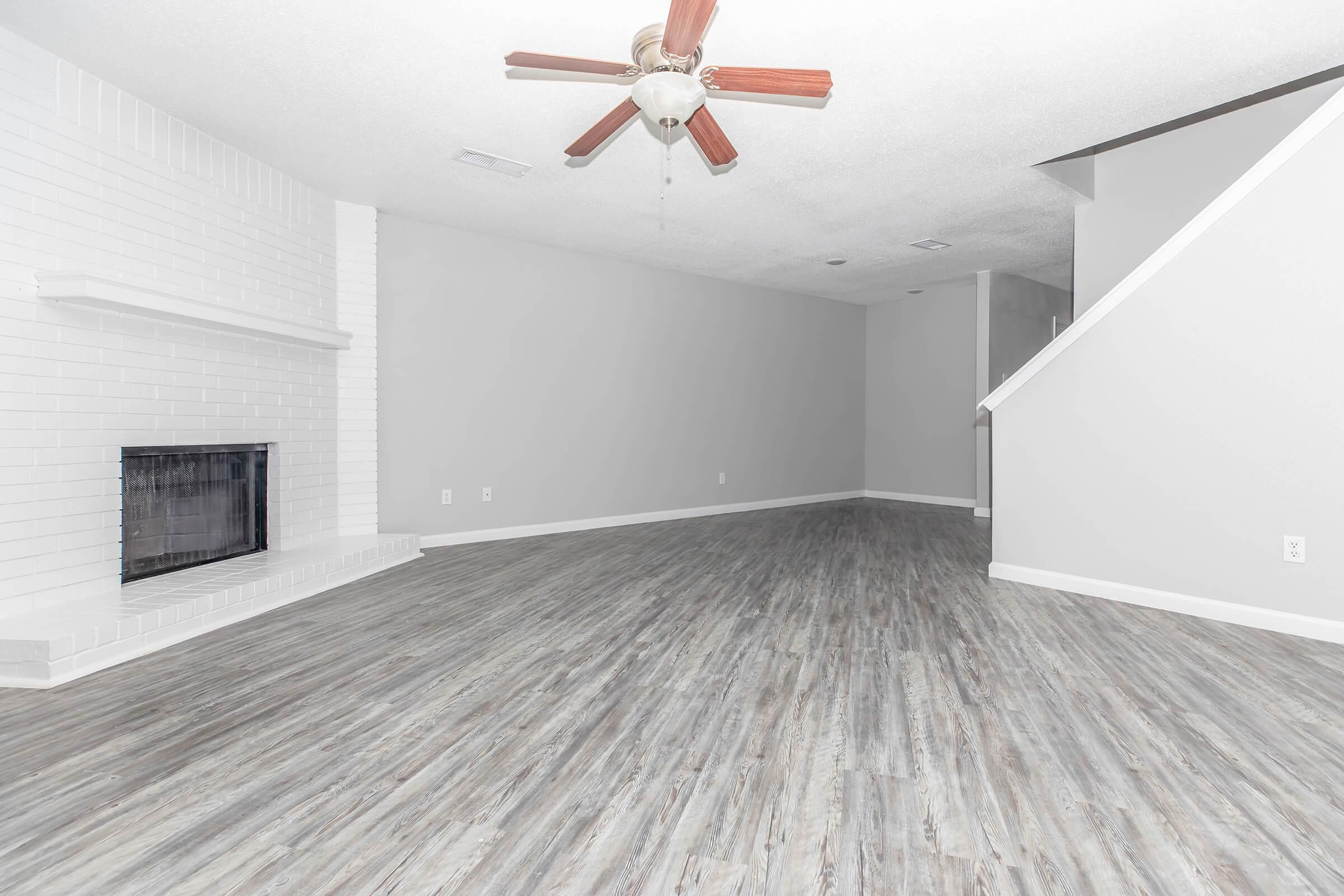 A spacious, empty living room with light gray walls and laminate flooring. There is a white brick fireplace on one wall, and a ceiling fan with wooden blades hangs above. Stairs lead to another level in the background, creating an open and airy atmosphere.