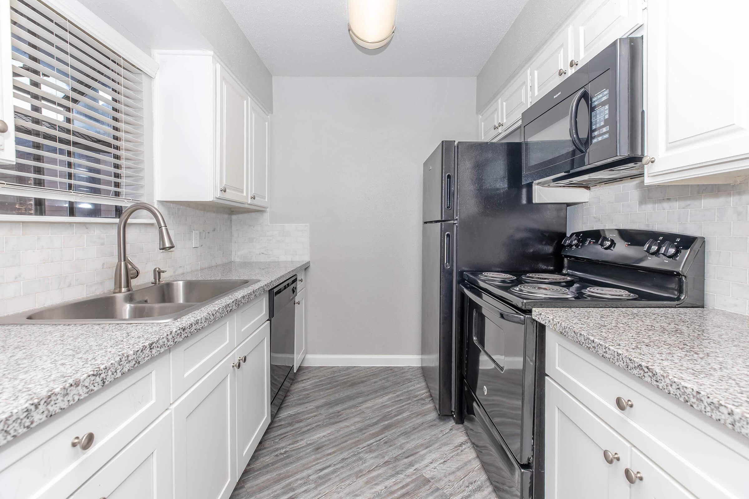A modern kitchen featuring white cabinetry, a gray granite countertop, and black appliances including a refrigerator and stove. The space has a window with blinds, providing natural light, and a stainless steel sink. The flooring is a light wood-like laminate.