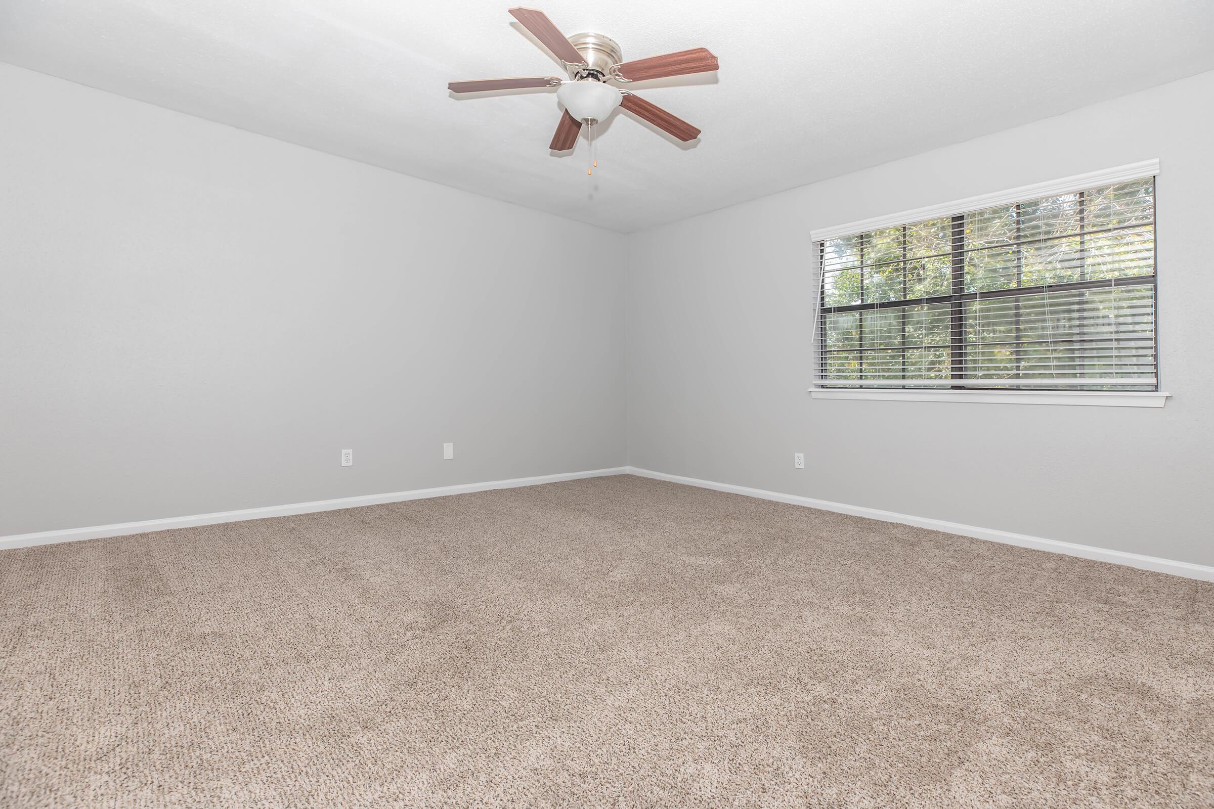 A spacious room with light gray walls and a ceiling fan. The floor is covered in soft, beige carpet. A window with blinds allows natural light to enter, and the overall atmosphere is airy and inviting. The room is uncluttered, providing a blank canvas for potential furnishings.