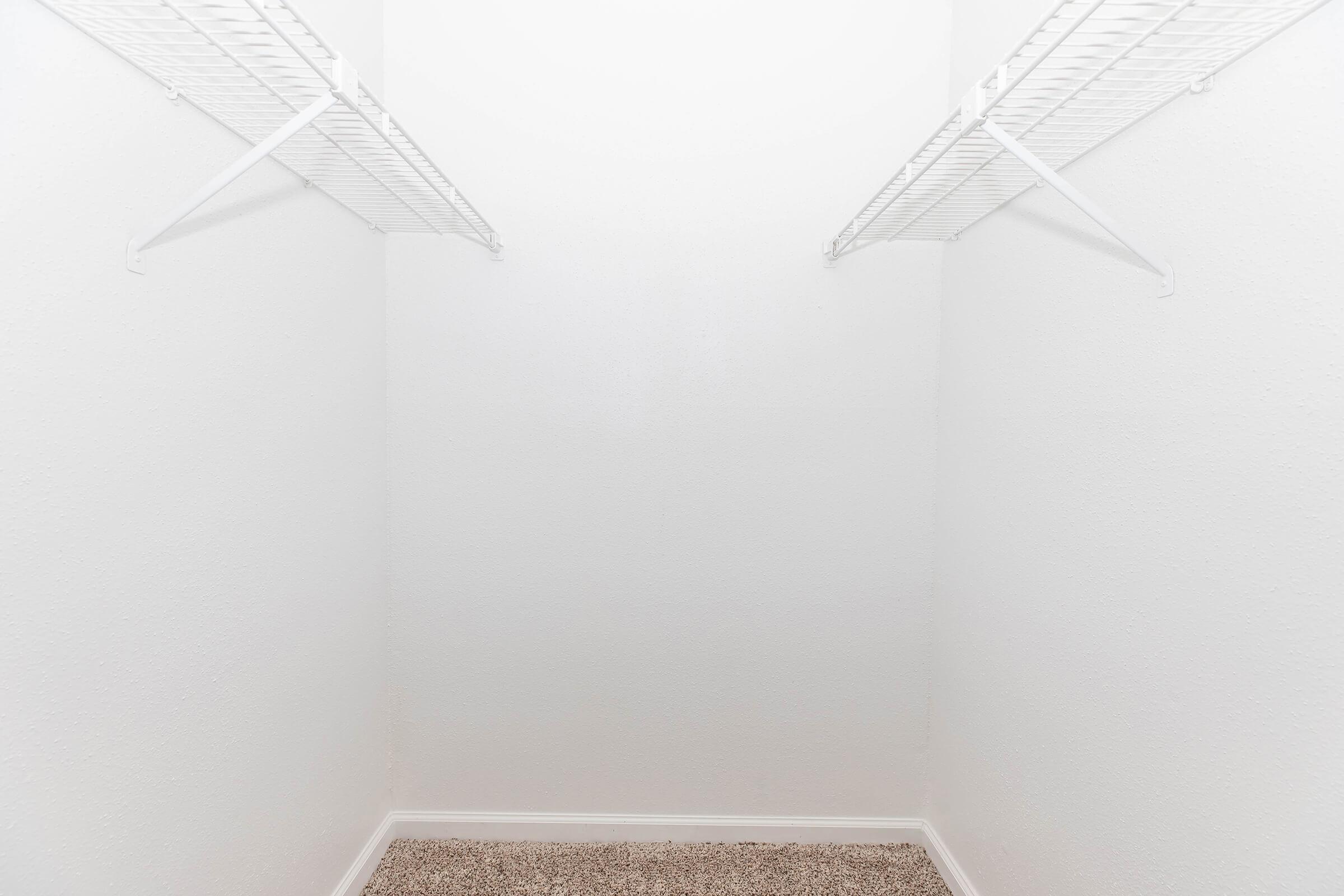 A clean, empty closet with white walls and a light-colored carpet. Two wire shelves are mounted on the side walls, providing storage space, while the floor remains clear and uncluttered. The overall appearance is bright and spacious.