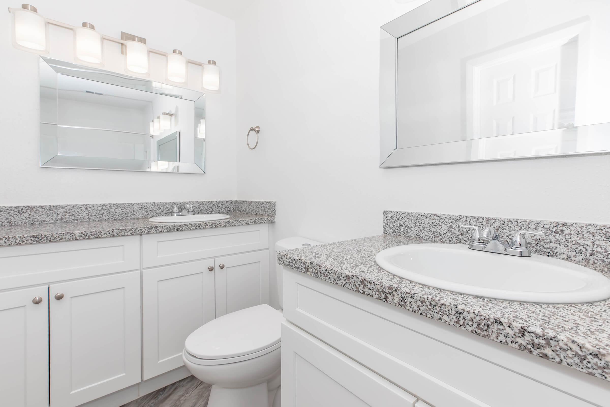 A modern bathroom featuring a white color scheme with granite countertops, a double sink vanity, and a large mirror. Bright lighting fixtures are mounted above the mirror, and a clean toilet is positioned beside the vanity. The floor has a subtle pattern, enhancing the sleek design of the space.