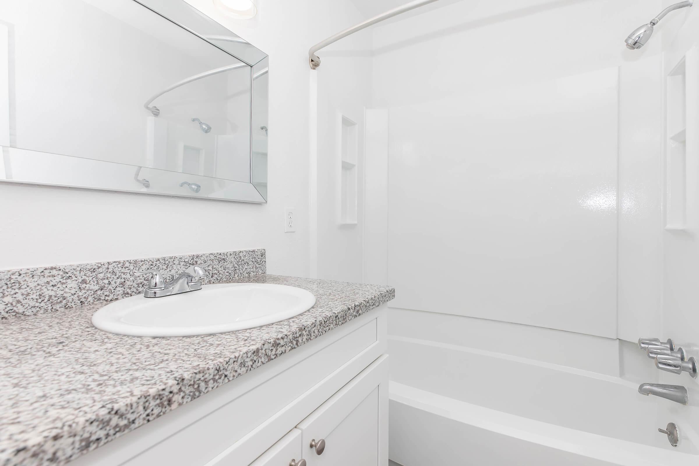 A clean and modern bathroom featuring a white bathtub with a curved shower rod, a granite countertop with a sink and faucet, and a wall-mounted mirror. The walls are painted white, creating a bright and airy atmosphere.