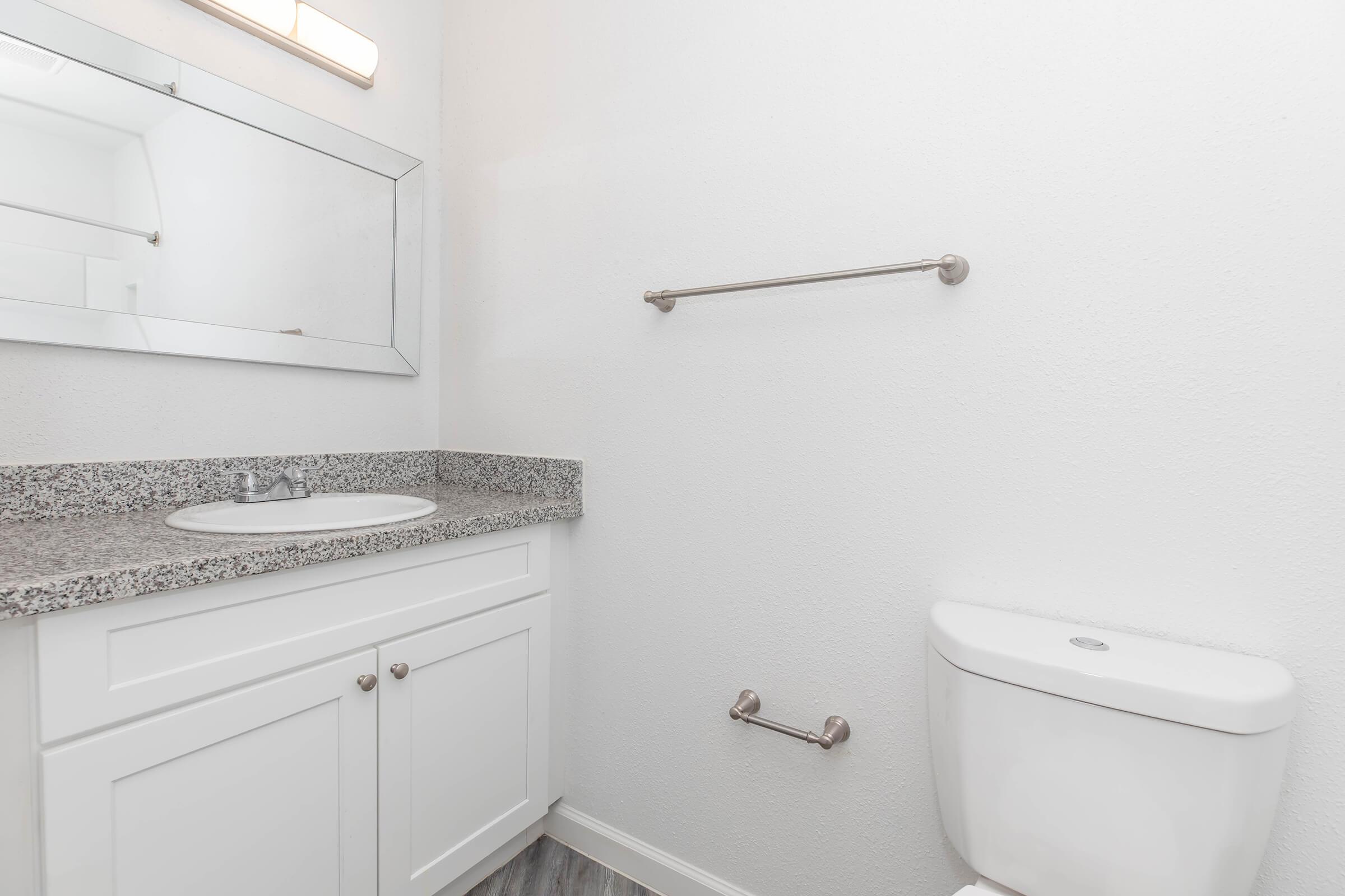A clean, modern bathroom featuring a white vanity with a granite countertop, a mirror above the sink, and a chrome towel bar on the wall. To the right, there is a white toilet. The walls are painted white, and the floor has a subtle pattern. A single light fixture is above the mirror.