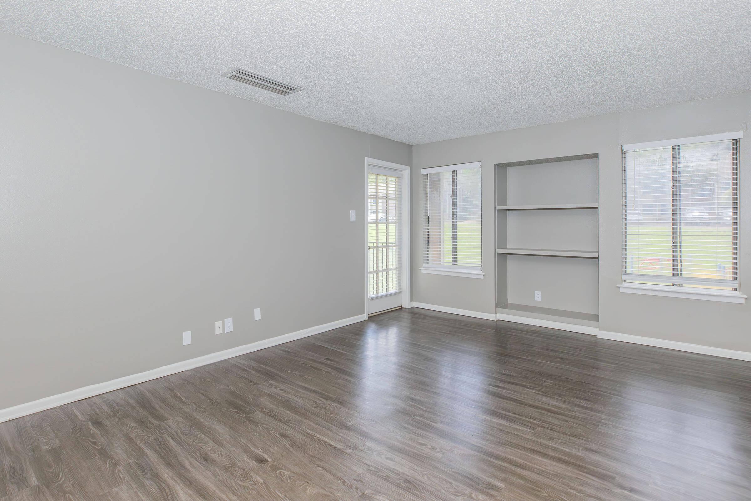 A spacious, empty living room featuring light gray walls, new hardwood flooring, and two large windows with blinds. There is a built-in shelf against one wall and a door leading outside. The room is well-lit, providing a bright and airy atmosphere.