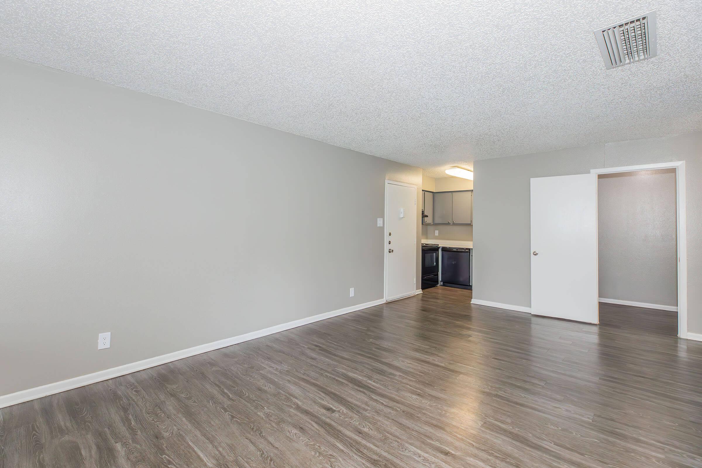 A spacious and empty living area with light gray walls and dark wood flooring. A doorway leads to a kitchen area with modern cabinetry in the background. The room features a textured ceiling and is well-lit, creating an inviting atmosphere for potential residents.