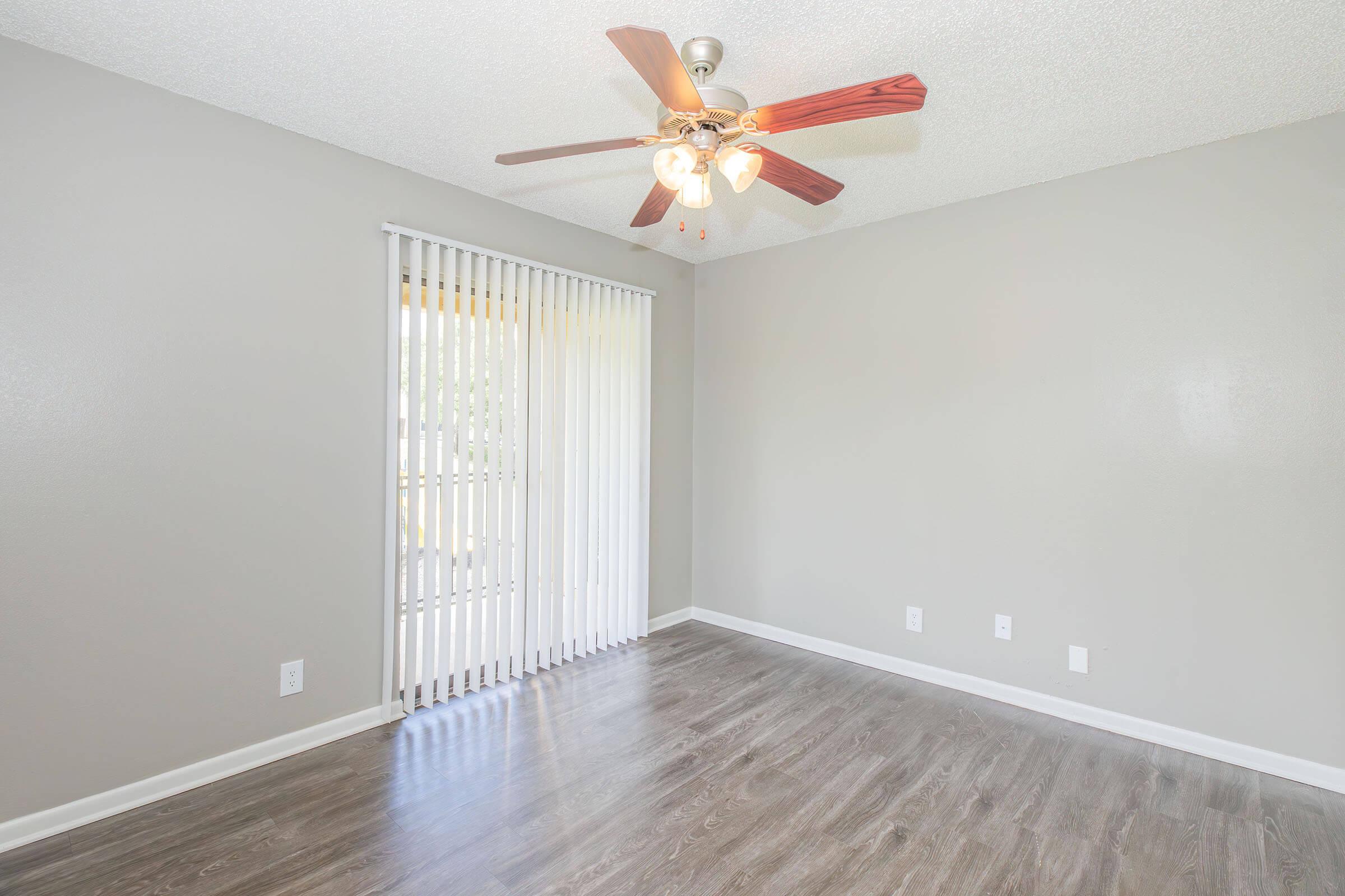 A spacious, empty room with light gray walls and a ceiling fan featuring wooden blades. There's a window with vertical blinds allowing natural light to enter, and the floor is covered with dark laminate flooring. The room has a clean and minimalist look, ideal for customization.