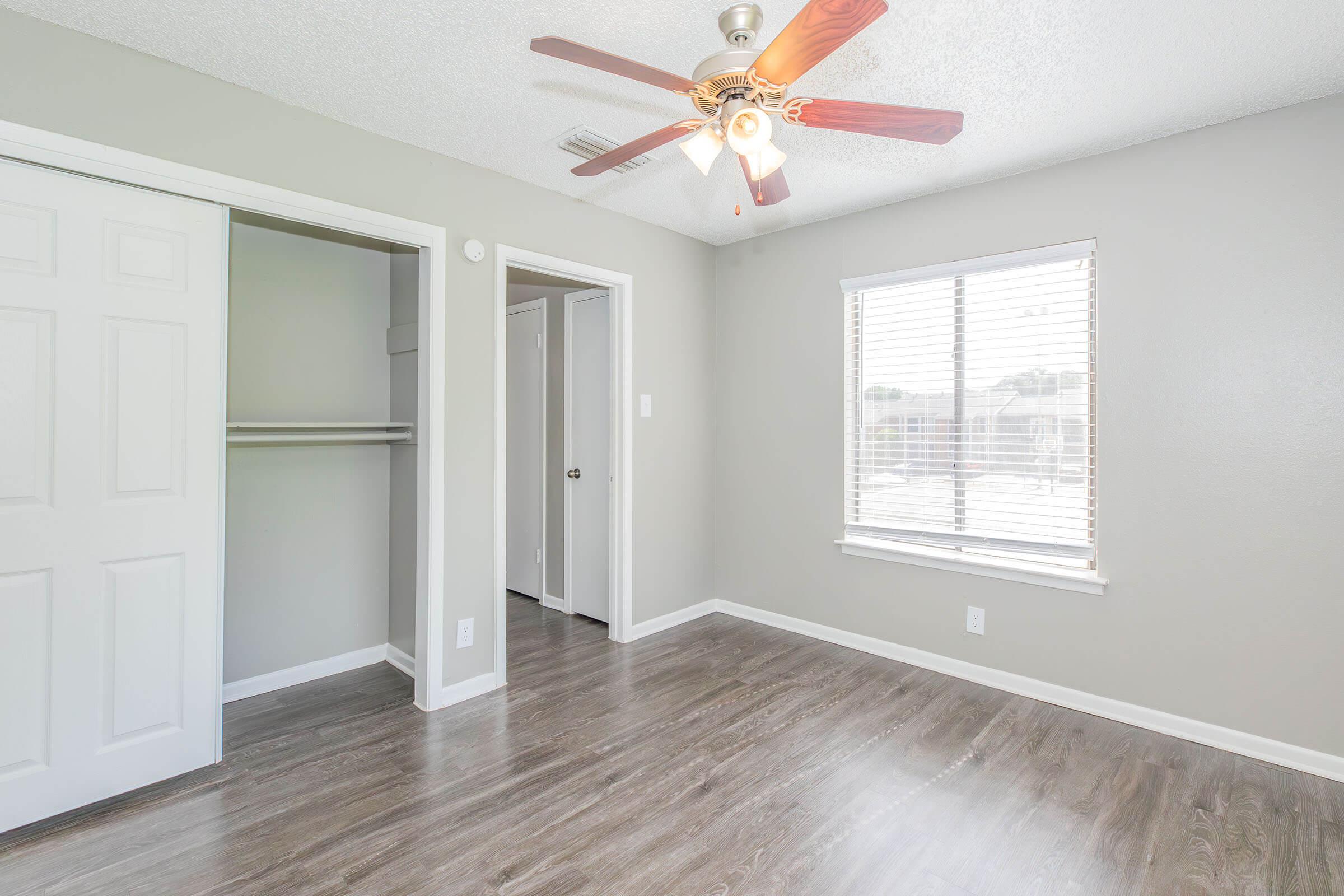 A bright, carpeted bedroom featuring a ceiling fan with wooden blades, a window with blinds, and a closet with sliding doors. The walls are painted a light gray, and the flooring is a light wood laminate. The room has a clean and modern look, with neutral colors and natural light streaming in from the window.