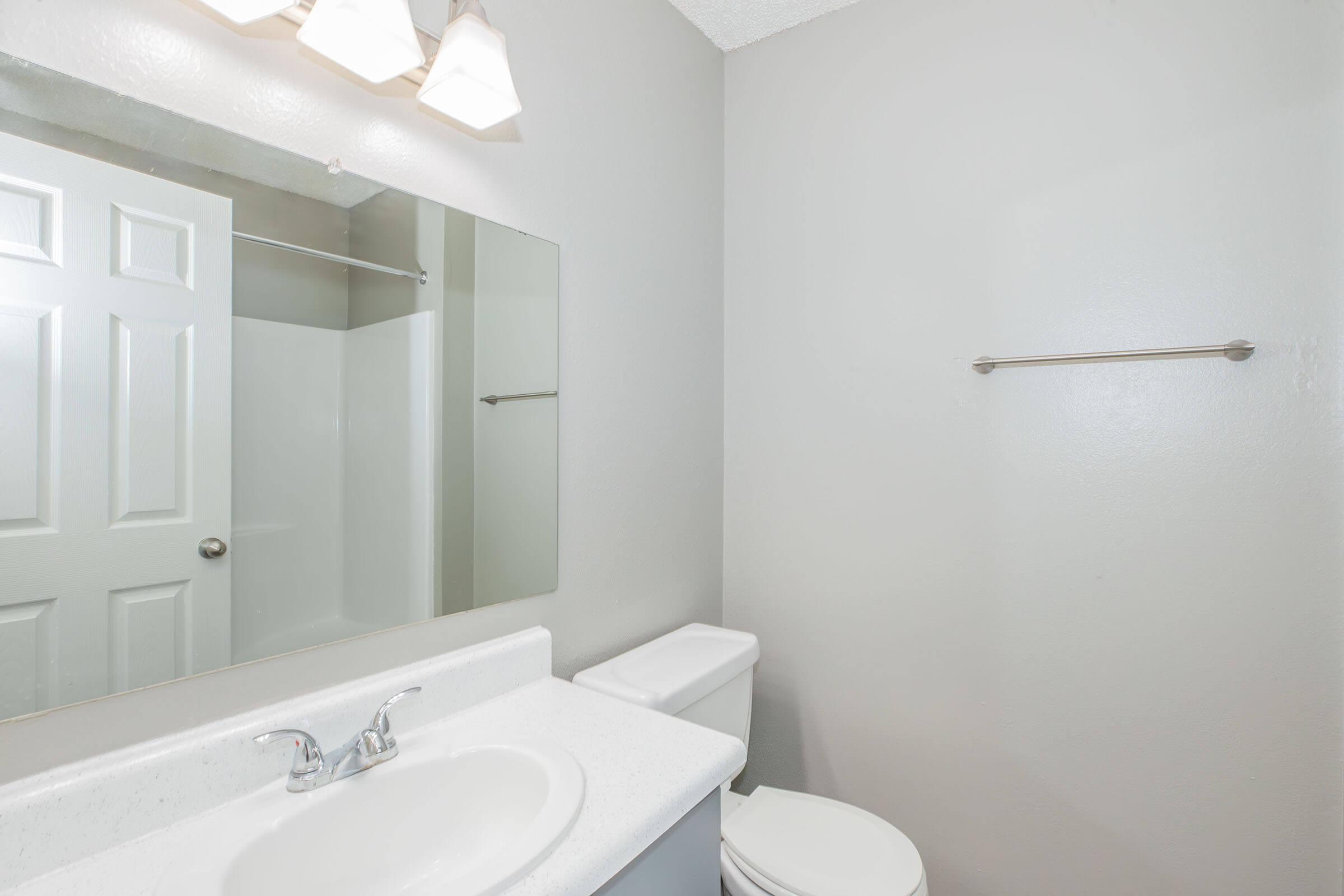 A clean, modern bathroom featuring a white sink with a faucet, a large mirror above the sink, a toilet, and a shower area with a sliding door. The walls are painted a light gray, and there is a simple towel bar on the wall to the right. The overall ambiance is bright and fresh.