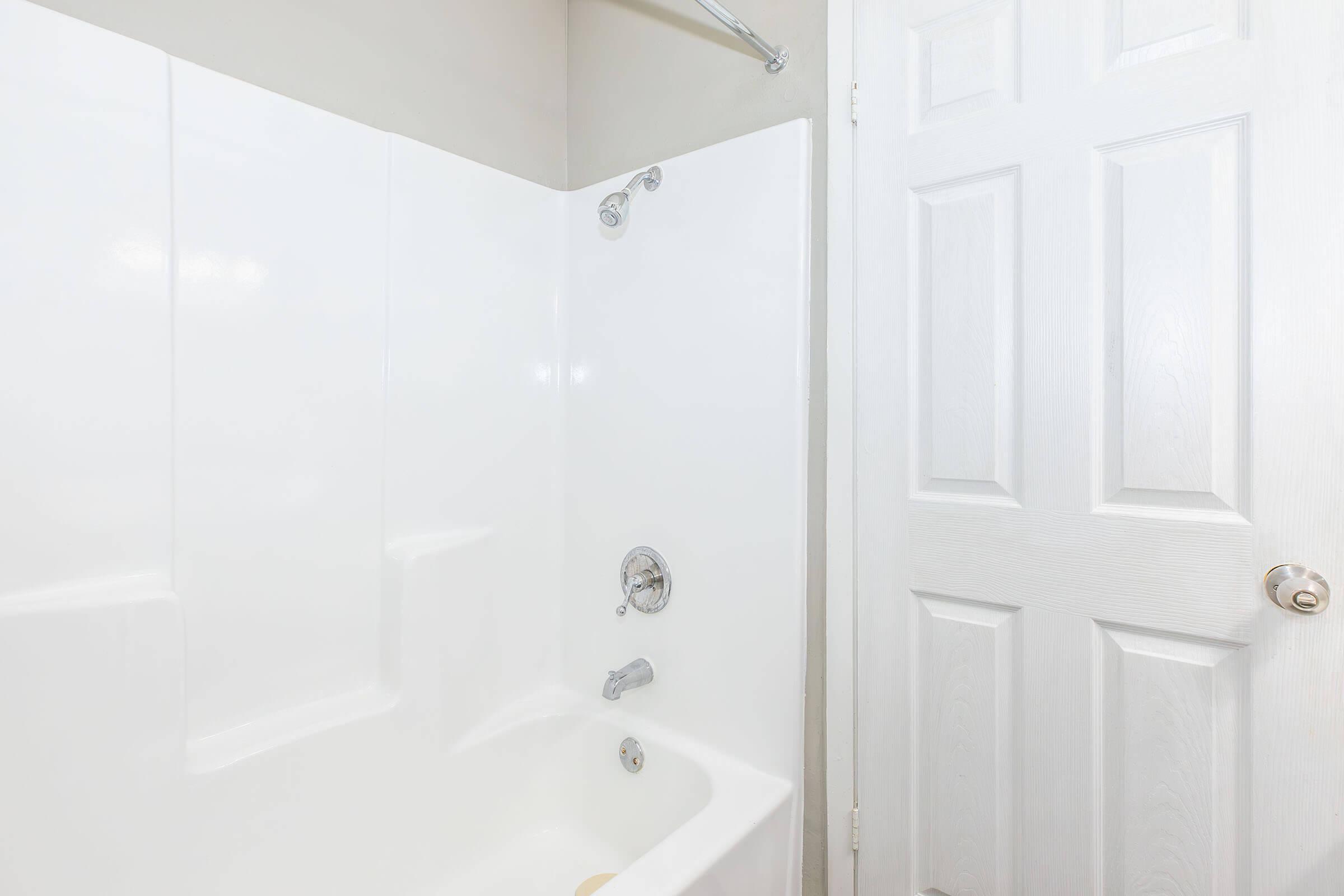 A clean, simple bathroom featuring a white bathtub with a shower head and faucet. There is a light-colored wall behind the tub, and a closed door with a round knob is visible to the right. The overall setting appears bright and well-maintained.