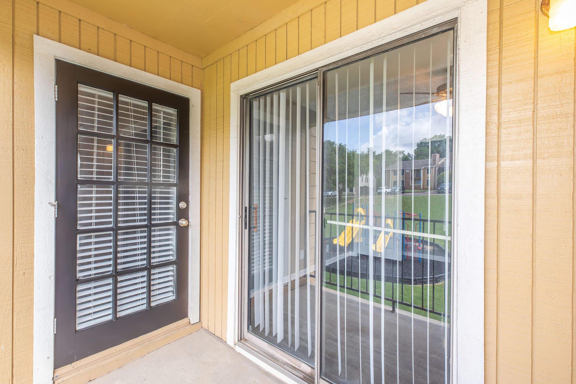 A view of an entrance with a brown door featuring glass panels. Adjacent is a sliding glass door with vertical blinds, revealing an outdoor area with grass and a playset in the background. The setting has a warm, inviting atmosphere, typical of residential porches.