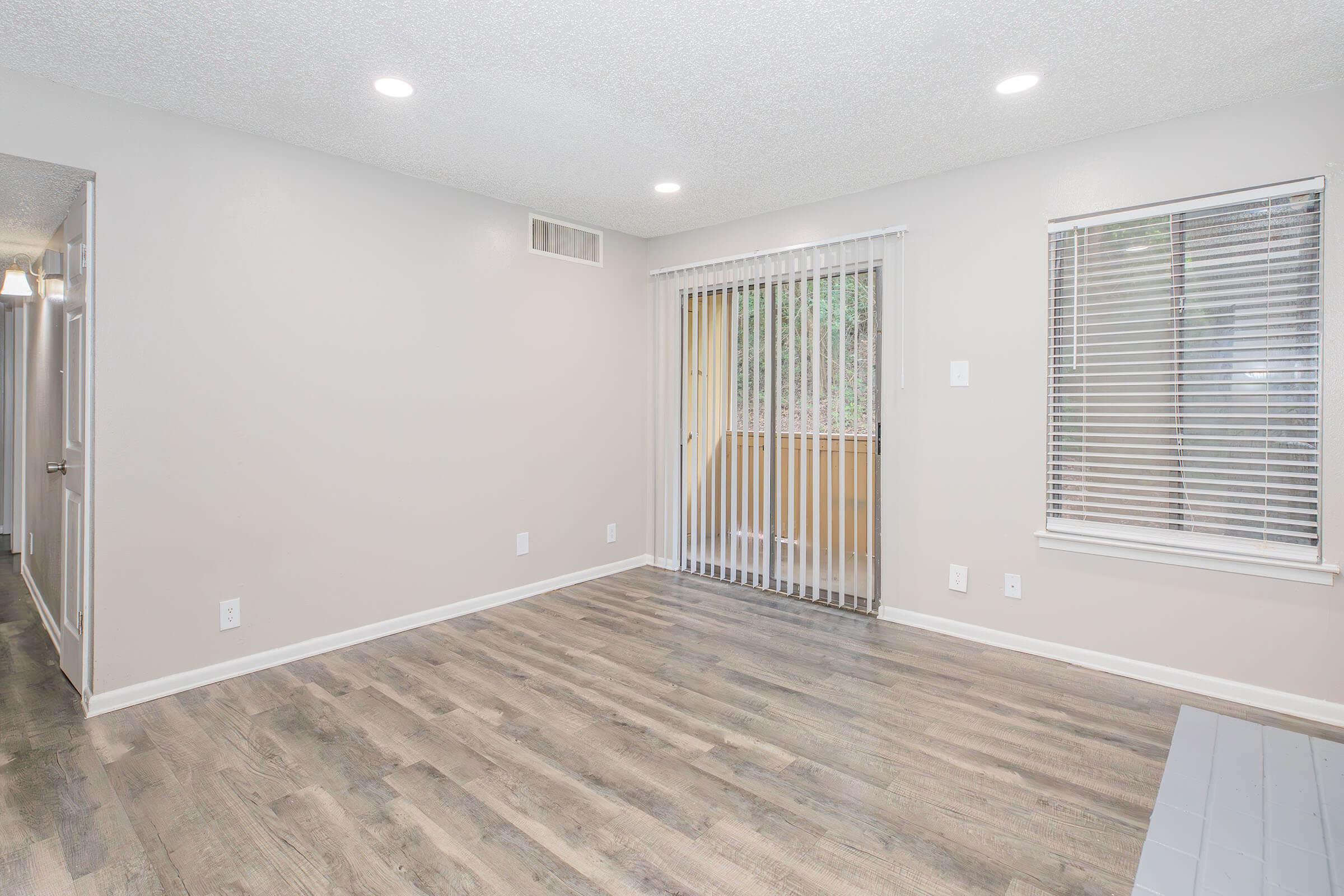 Spacious, empty room with light-colored walls and laminate flooring. Features large windows with vertical blinds allowing natural light, and a sliding door leading to an outdoor area. The room is well-lit with overhead lights, creating a bright and open atmosphere. Ideal for living or dining space.