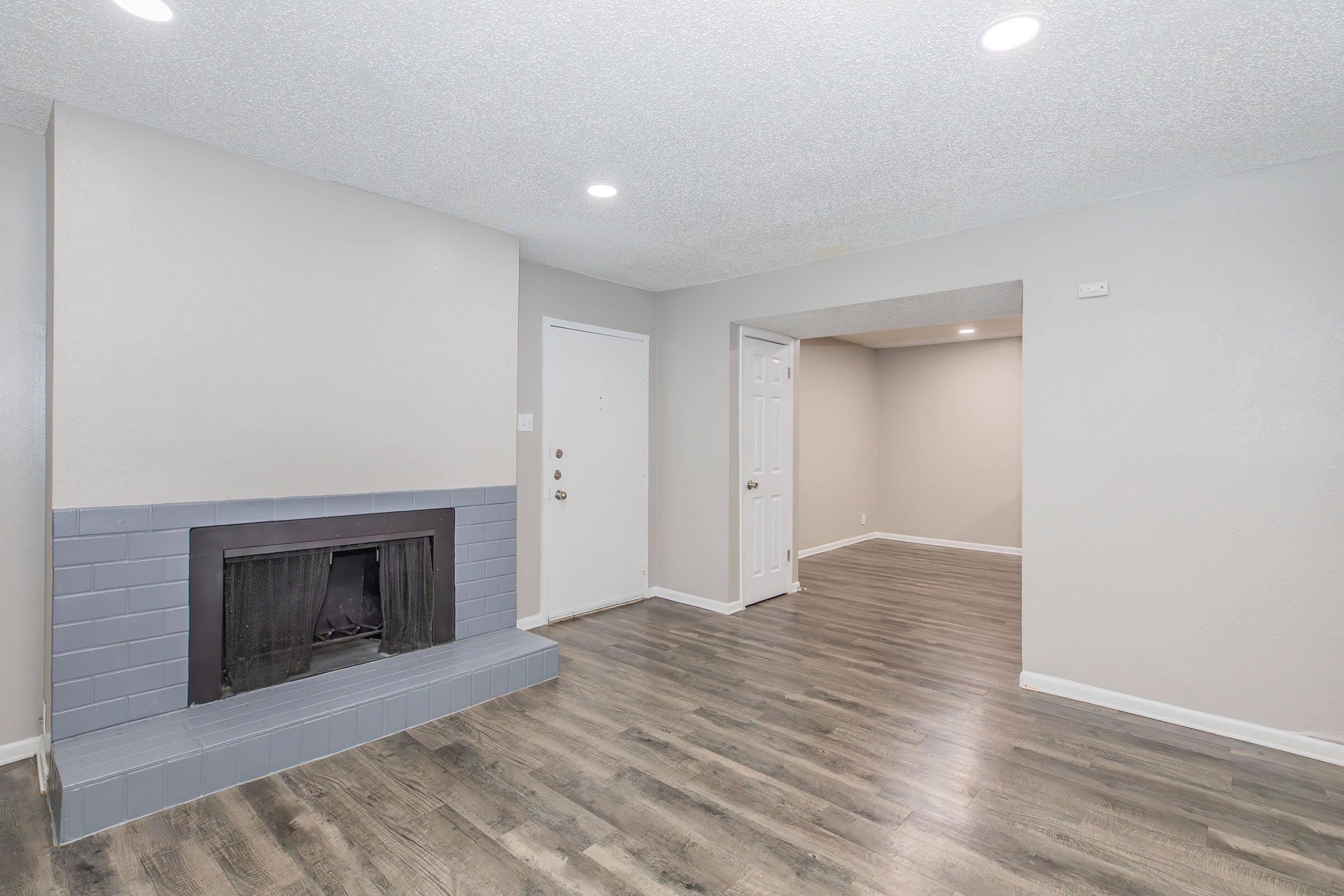 A spacious living room featuring a gray fireplace on the left, light-colored walls, and laminate flooring. The entrance door is on the left, and there is an open area leading to another room in the background, illuminated by ceiling lights.