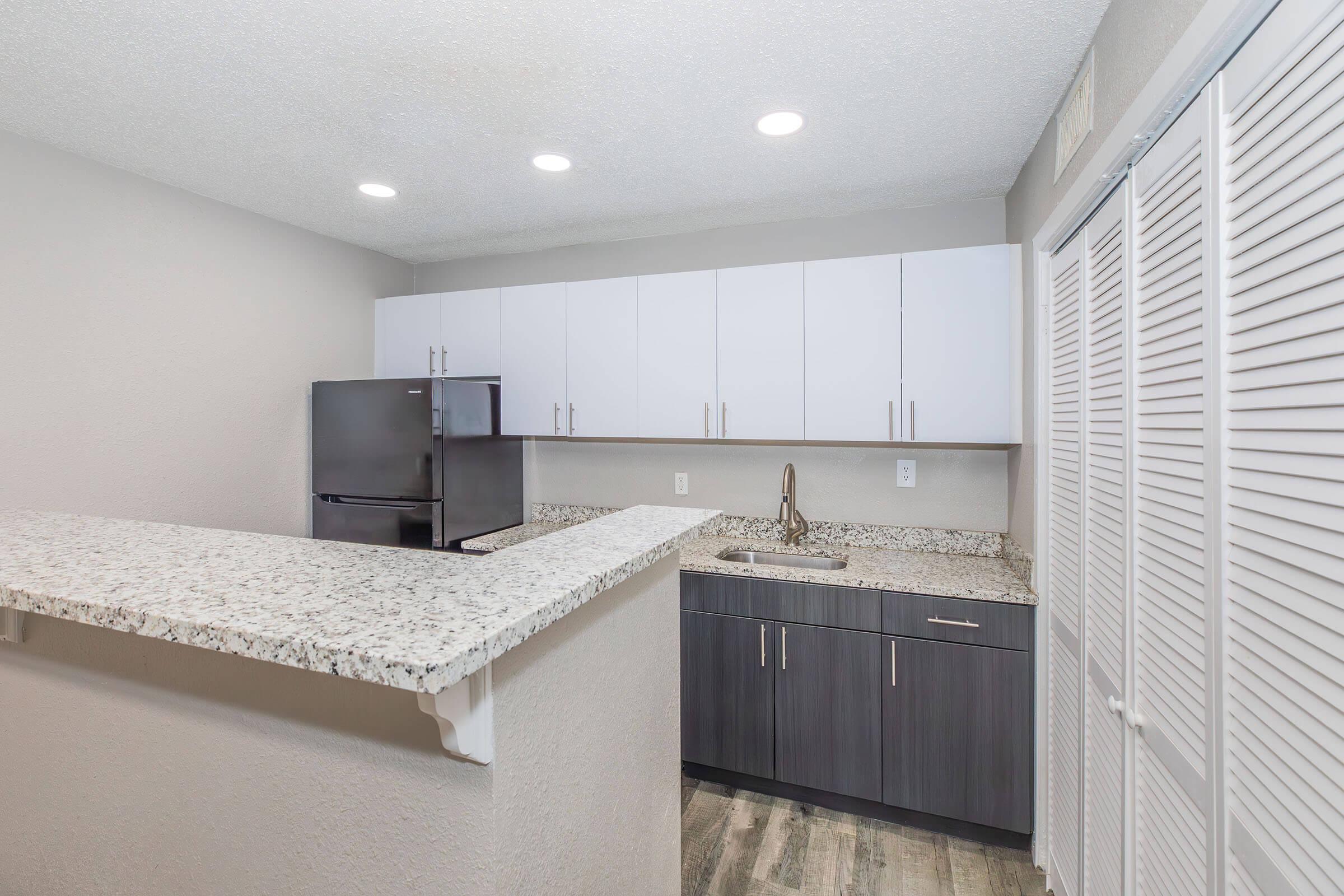 Modern kitchen featuring white and dark wood cabinetry, a granite countertop, and stainless steel appliances, including a refrigerator. The layout is open with recessed lighting, and there is a bar area at the countertop, creating a functional and stylish space.