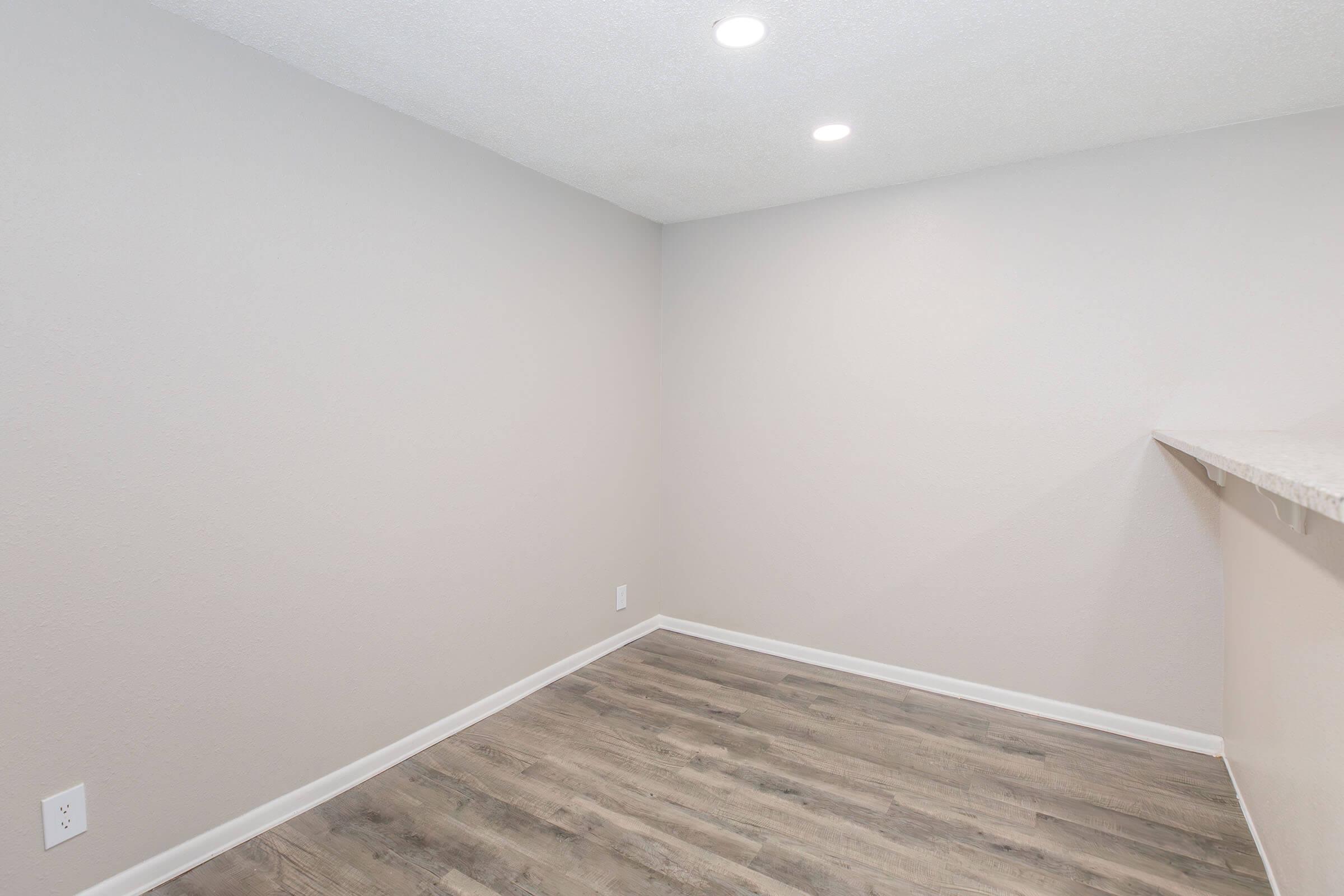 Empty room with a light gray wall, featuring recessed ceiling lights. The floor has a wood-like laminate finish, and there is a small opening leading to a countertop area, possibly a kitchen bar. The overall ambiance is bright and minimalistic.
