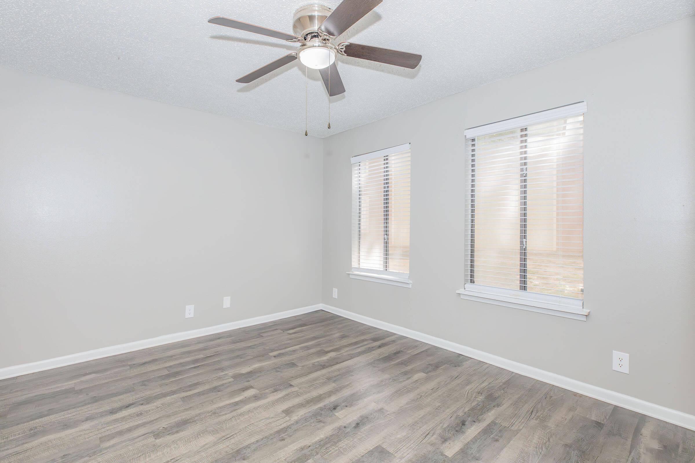 A bright and airy room featuring light gray walls, two windows with white blinds, and a modern ceiling fan. The floor is made of light wood laminate, creating a cozy atmosphere in the space. A single electrical outlet is visible on the wall.