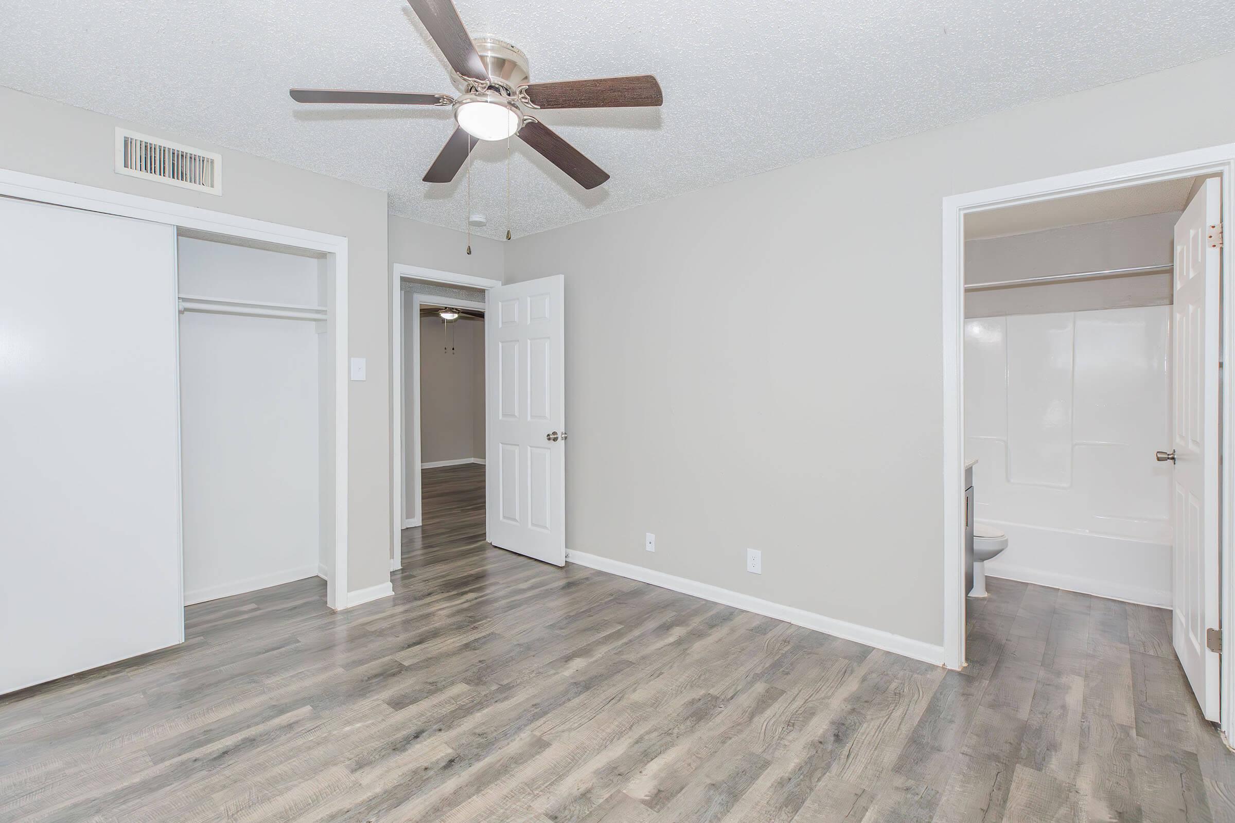A spacious room with a ceiling fan, light-colored walls, and laminate flooring. There are two openings: one leads to a bathroom with a bathtub, and the other features a closet. The room is well-lit, providing a welcoming ambiance.