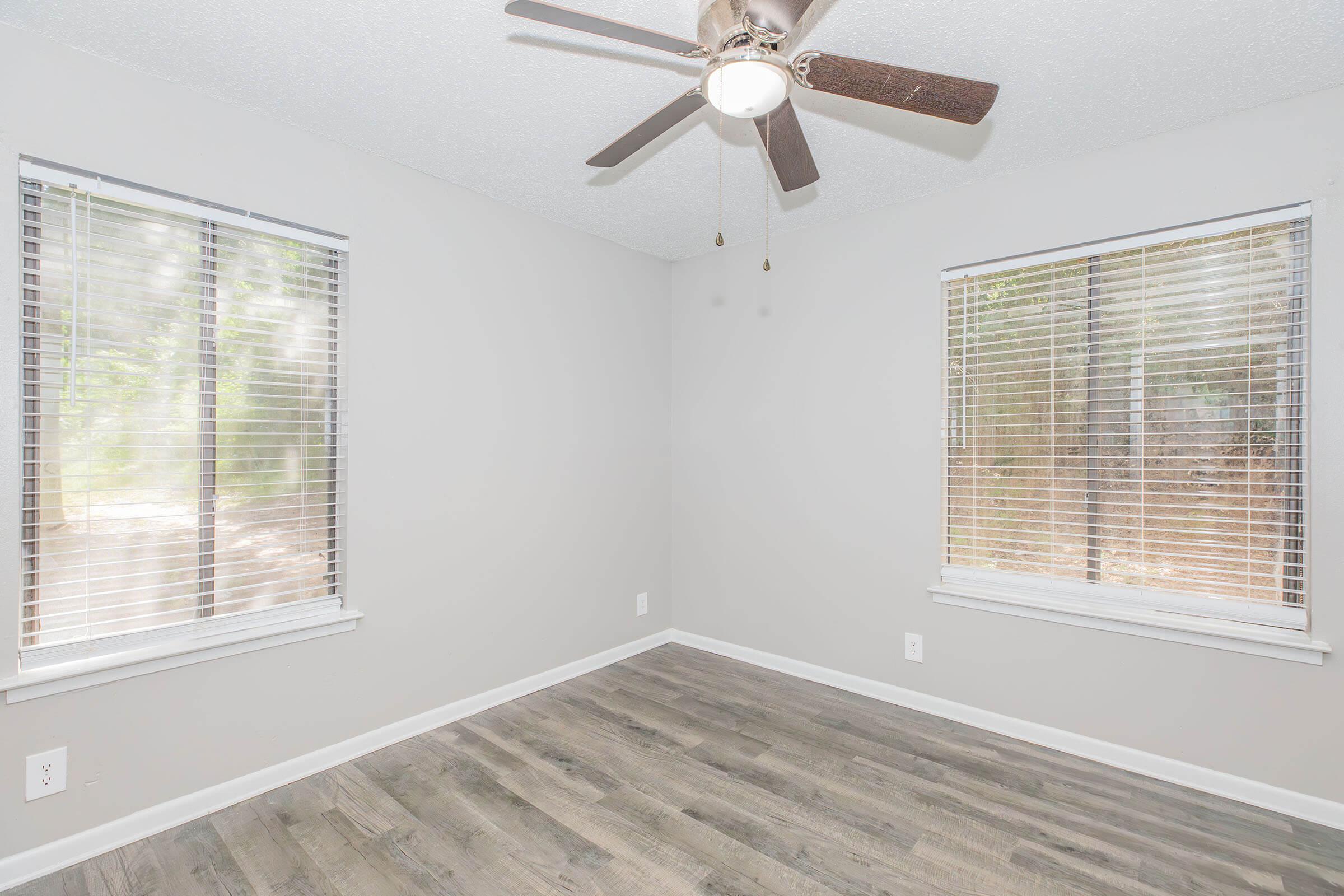 A well-lit, empty room with two large windows featuring blinds, a ceiling fan with light fixture, and light-colored walls. The flooring is a modern, wood-like finish, creating a clean and airy atmosphere. The view outside the windows shows a natural, wooded area, adding a touch of greenery to the space.