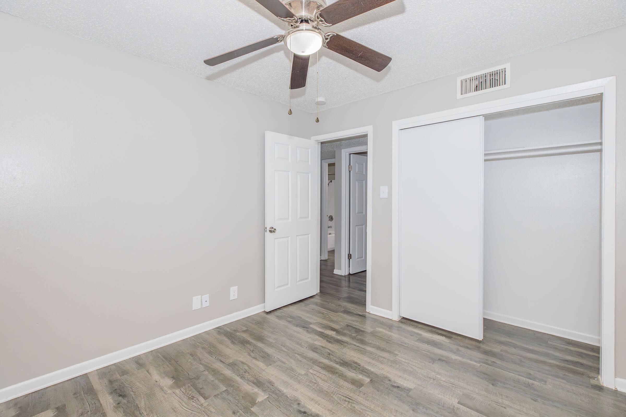 A well-lit, neutral-colored room featuring a ceiling fan, light wood-like flooring, and two doors leading to other spaces. One door opens to a closet. The walls are painted in a light shade, creating a clean and spacious atmosphere.