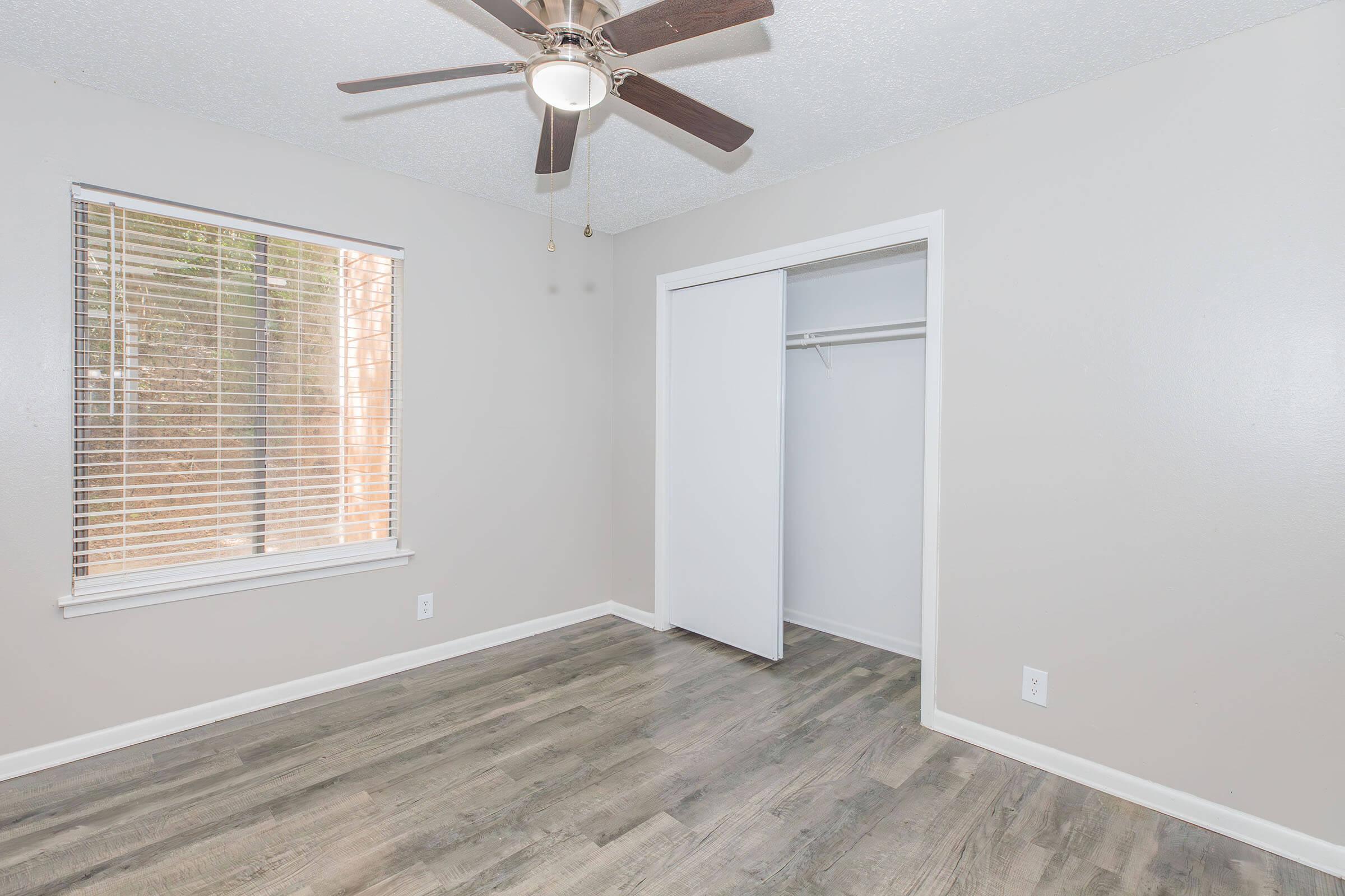 A well-lit room featuring a ceiling fan, a window with blinds, and light-colored walls. The floor is covered with gray laminate, and there is a white closet with sliding doors. The overall atmosphere is clean and minimalistic, ideal for a bedroom or office space.