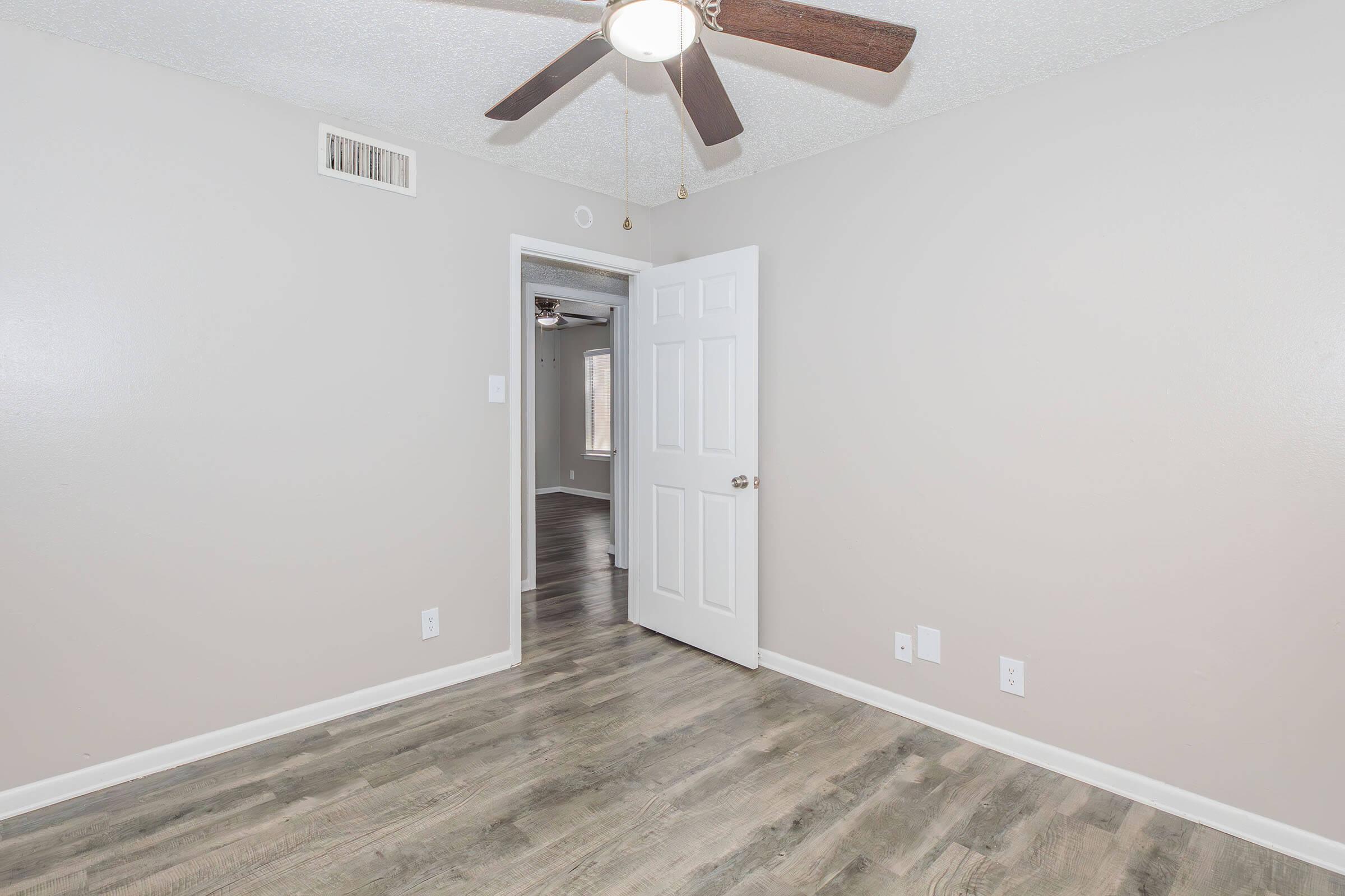 A well-lit, empty room featuring light gray walls and a modern ceiling fan. A white door is partially open, leading to another room. The floor is covered in a light wood laminate, creating a clean and inviting atmosphere.