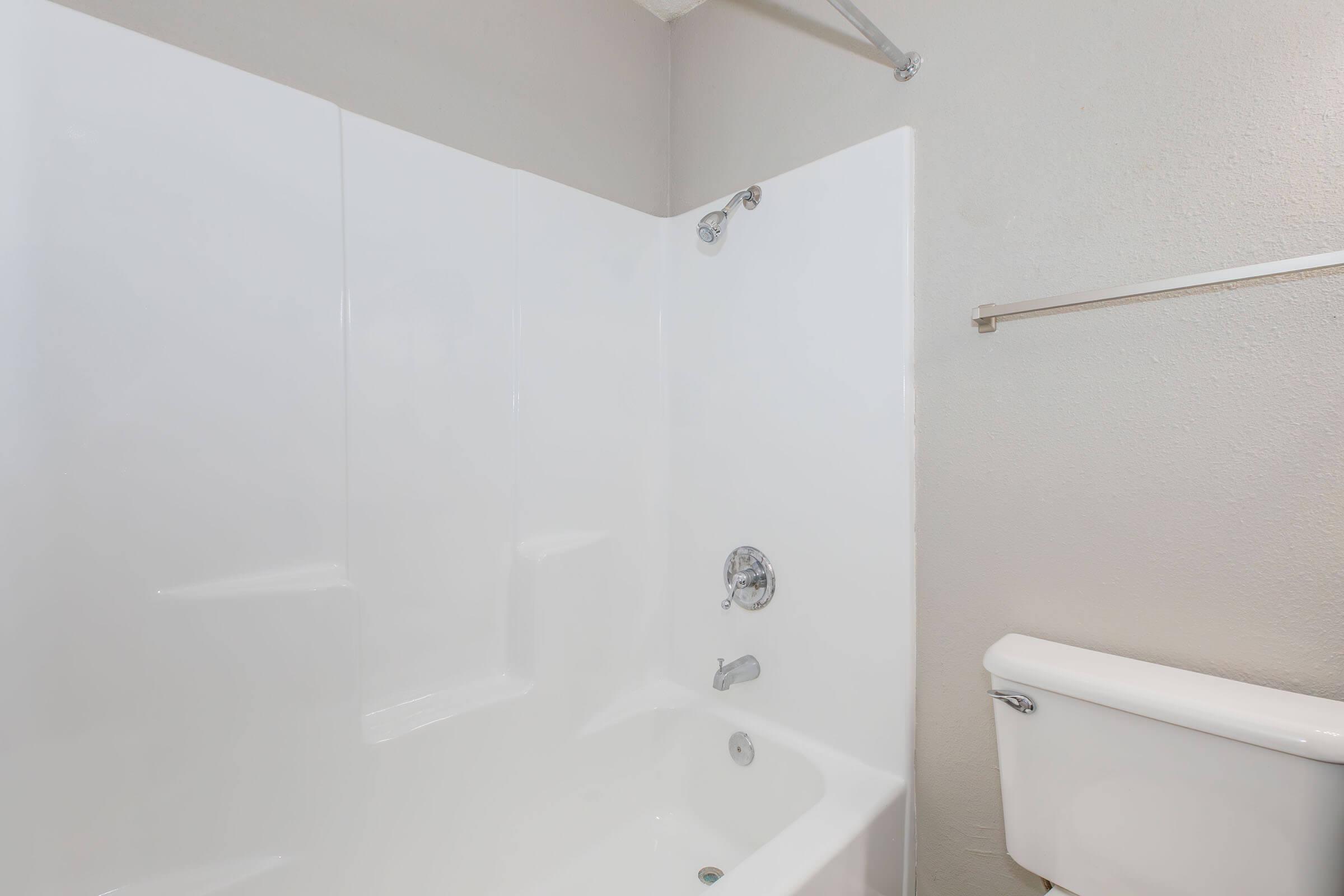 A clean, modern bathroom featuring a white tub and shower combo with a chrome faucet and showerhead. The wall is smooth and plain, and there is a toilet next to the tub. The overall aesthetic is minimalistic and bright, with neutral tones.
