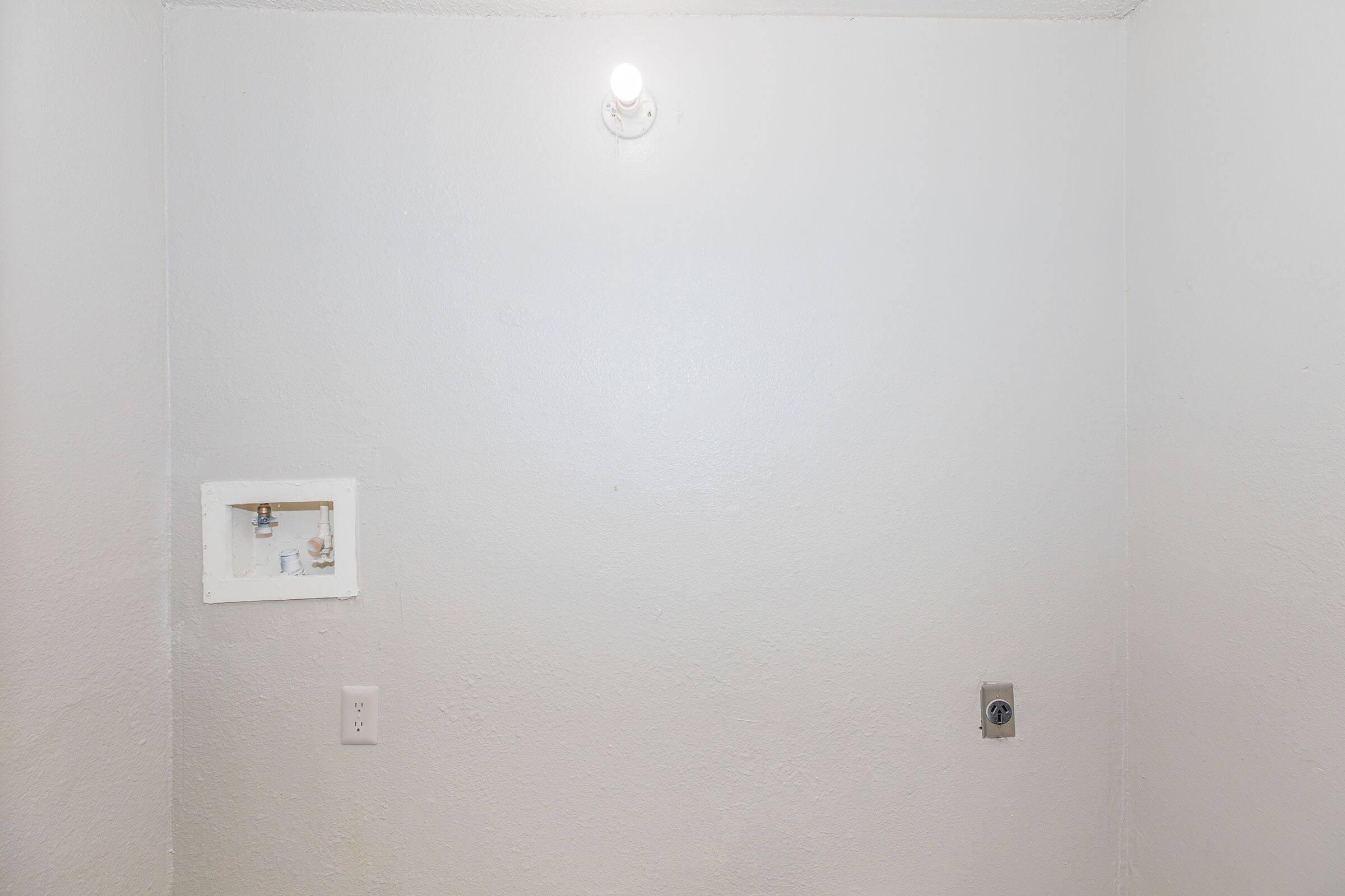 A plain, empty room with white walls. There is a small electrical panel on the left side and a light fixture on the ceiling illuminating the space. The room has no furniture or decorations, giving it a stark, minimalistic appearance.