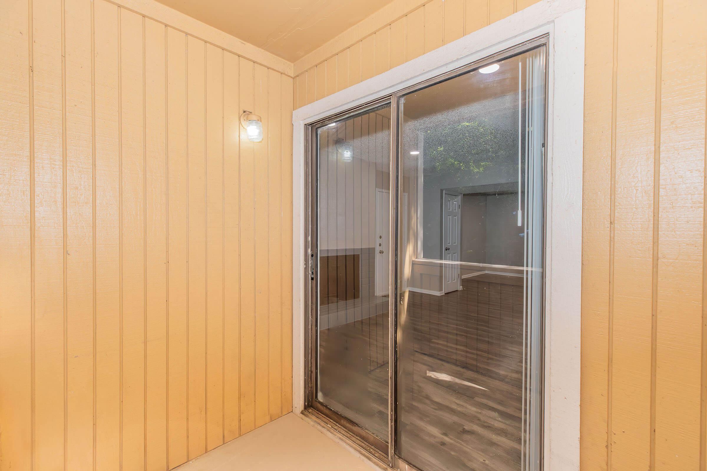 Exterior view of a sliding glass door leading into a room with light-colored walls and hardwood flooring. The surrounding walls are painted a soft yellow. The area is well-lit, with a light fixture on the wall to the left. The door's window provides a glimpse into the interior space beyond.