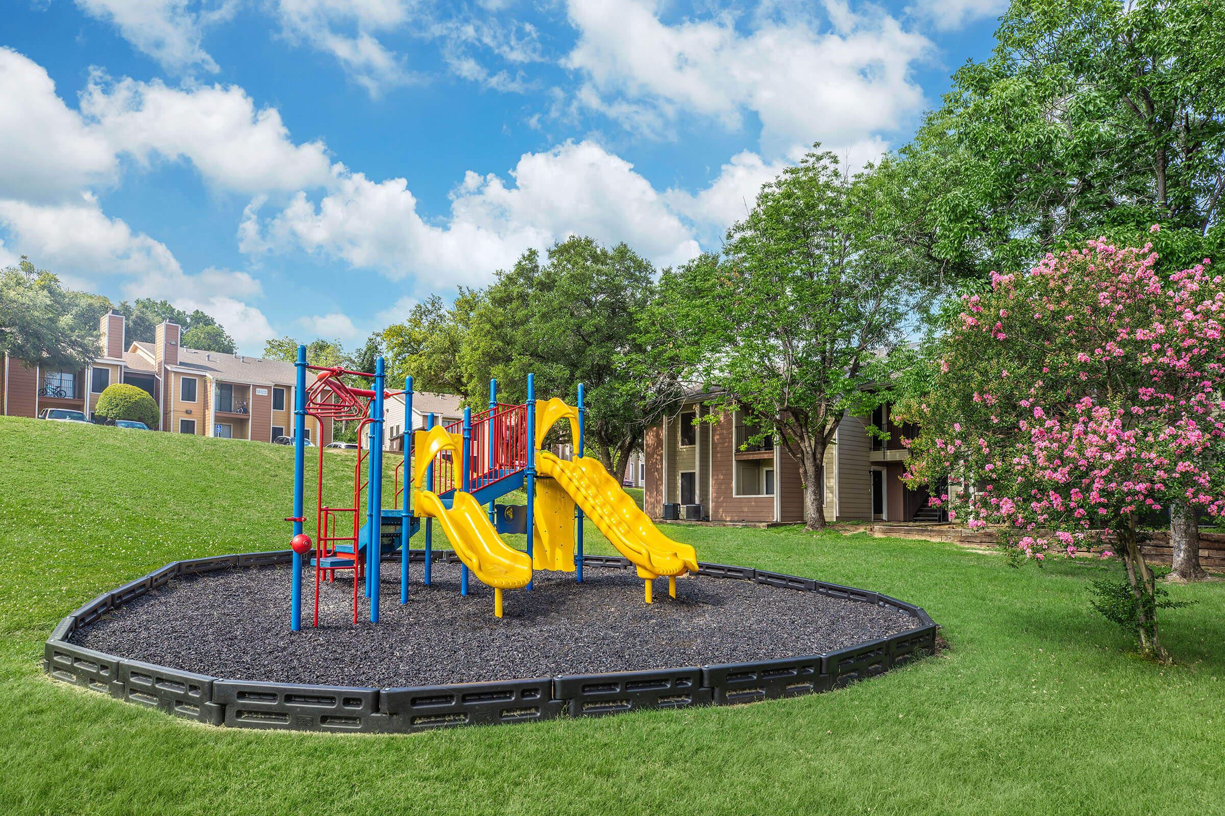A colorful playground featuring a yellow and blue slide, surrounded by green grass and trees. The area is bordered by black rubber matting, with an apartment complex visible in the background under a partly cloudy sky.