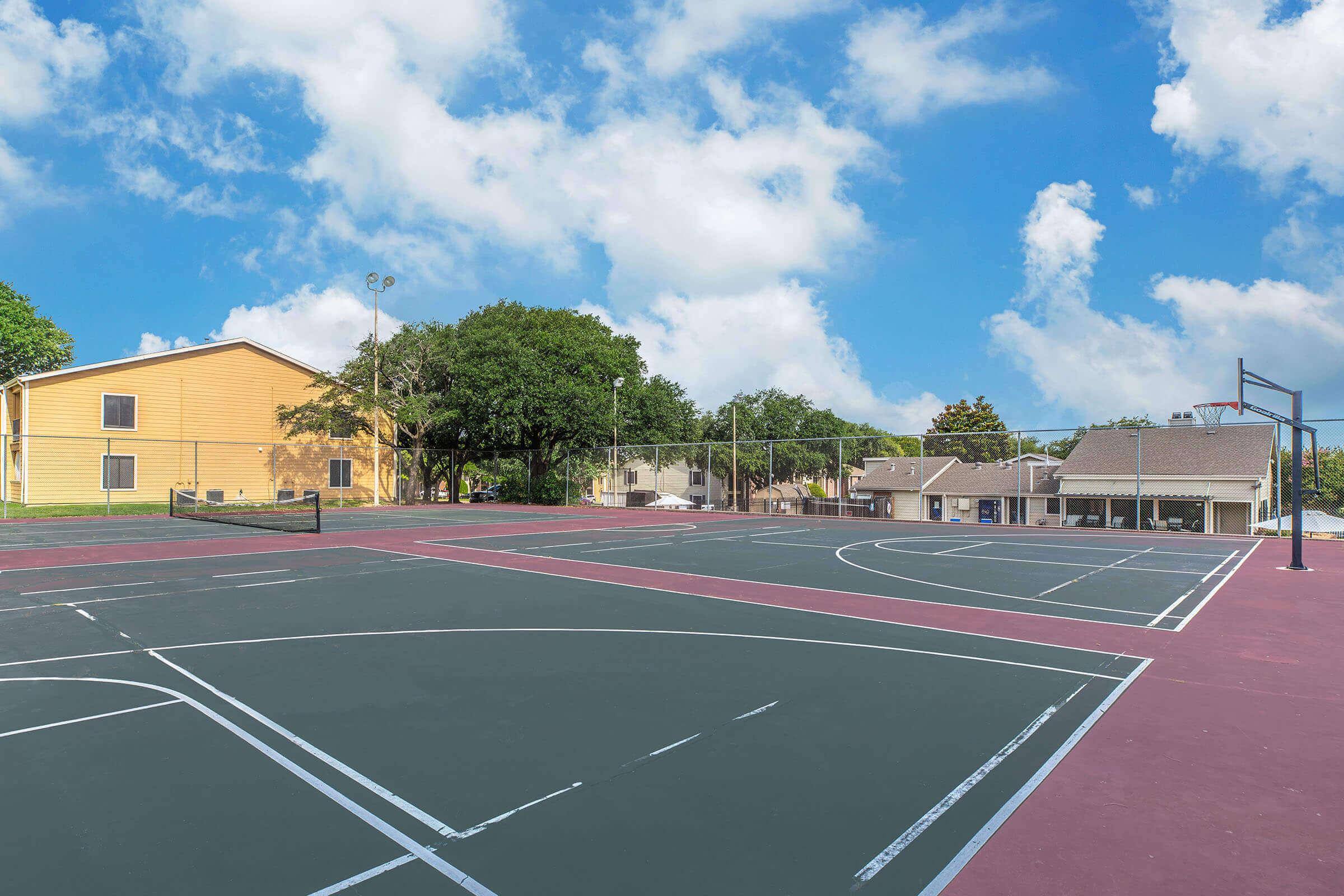 A basketball court featuring dark green and maroon painted surfaces, surrounded by trees and buildings. The sky is bright with fluffy clouds, and the area is well-maintained, inviting for outdoor sports activities.