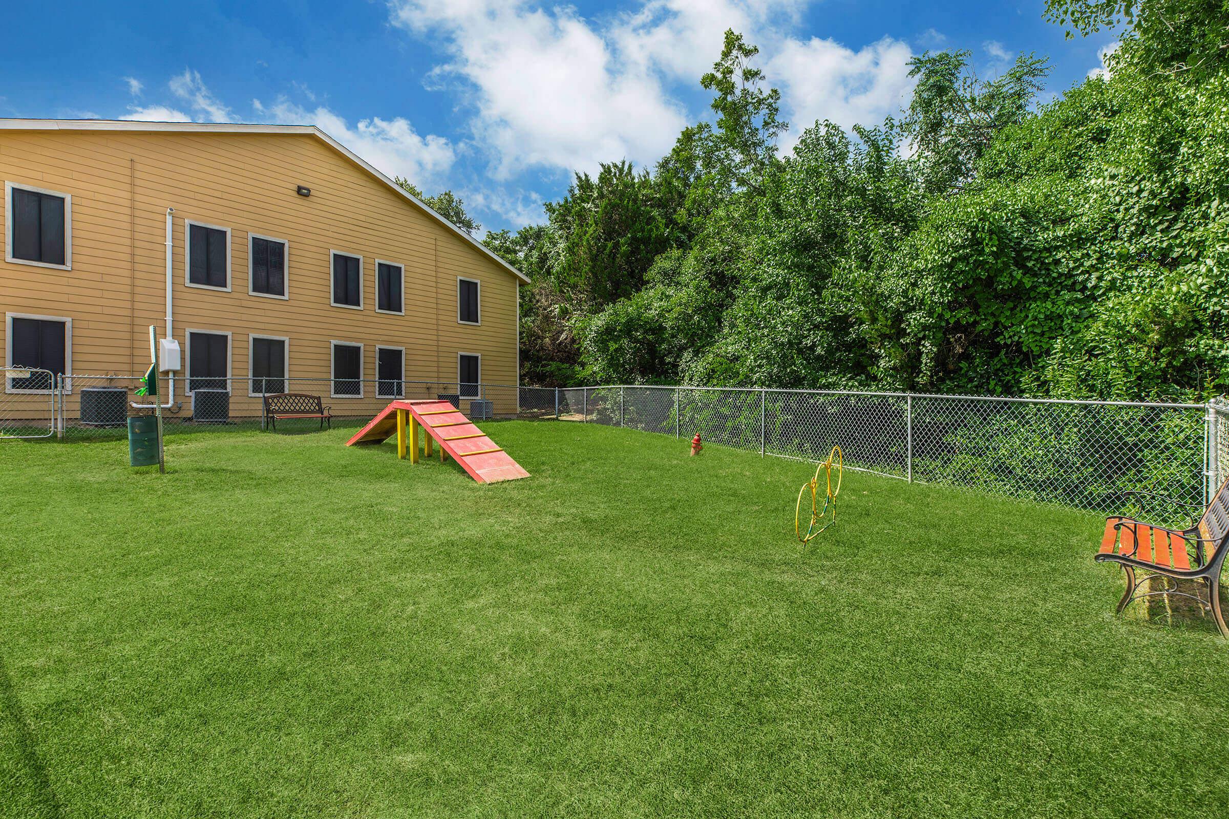A grassy dog park featuring agility equipment, including a ramp and hoops, enclosed by a chain-link fence. In the background, there is a yellow building with several windows and lush greenery surrounding the area, under a partly cloudy blue sky. A bench is visible in the foreground.
