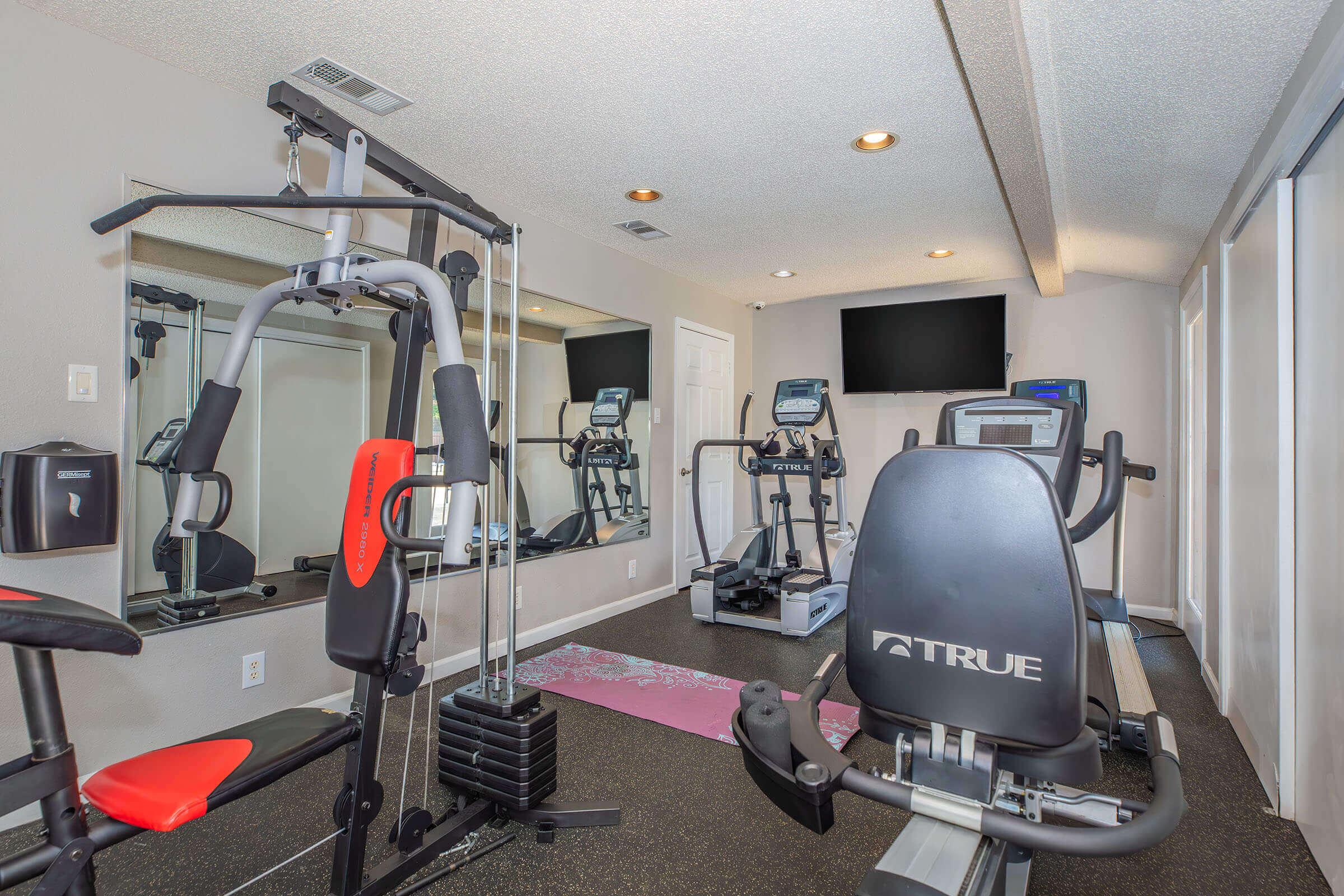 A home gym space featuring various exercise equipment, including a multi-use machine, a stair stepper, and an elliptical machine. The room has large mirrors and natural light from windows, creating a bright and motivating atmosphere for workouts.