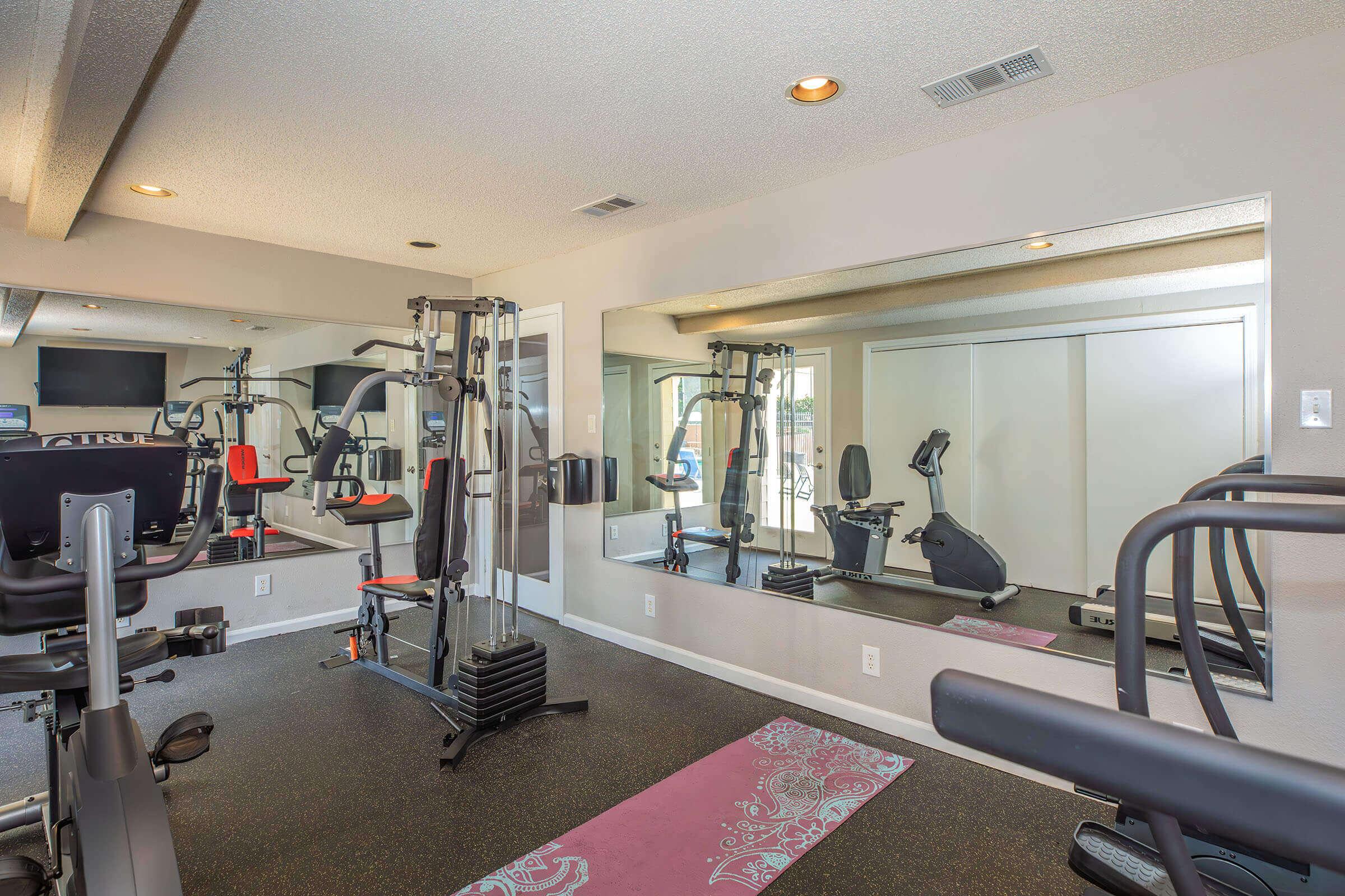 A modern gym with exercise equipment including a multi-gym machine, a stationary bike, and a treadmill. The space features a mirror on one wall and a yoga mat on the floor, enhancing the workout environment. Bright lighting and a clean design create an inviting atmosphere for fitness activities.